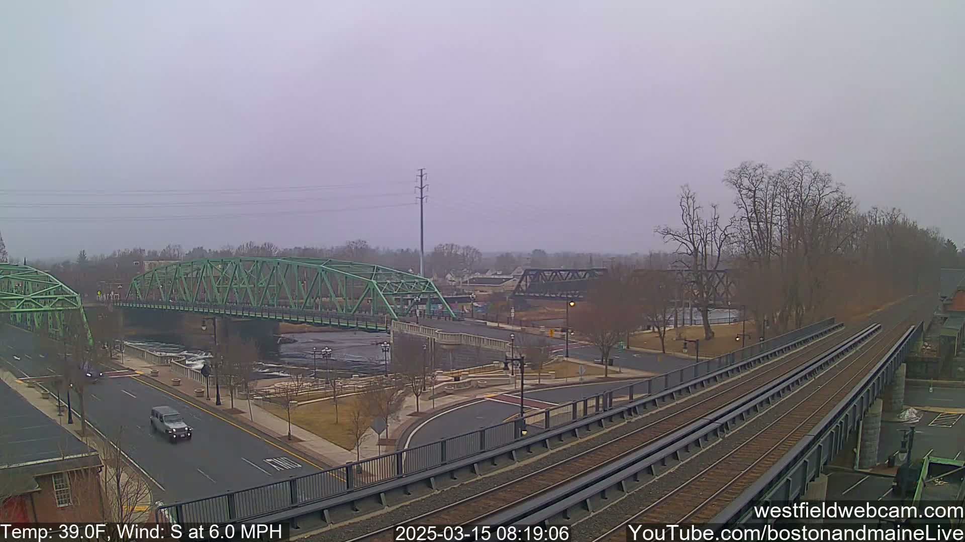 A view of a river with two bridges, one green and one black, under an overcast sky, with train tracks in the foreground.