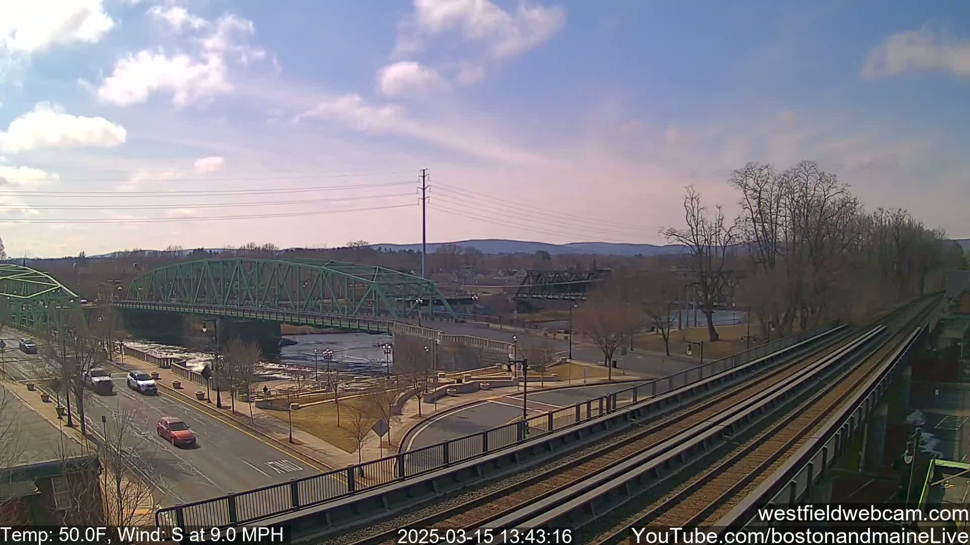 A green bridge spans a river, with cars driving on a road below and train tracks visible in the foreground under a partly cloudy sky, a temperature of 50°F, and a south wind at 9.0 mph.