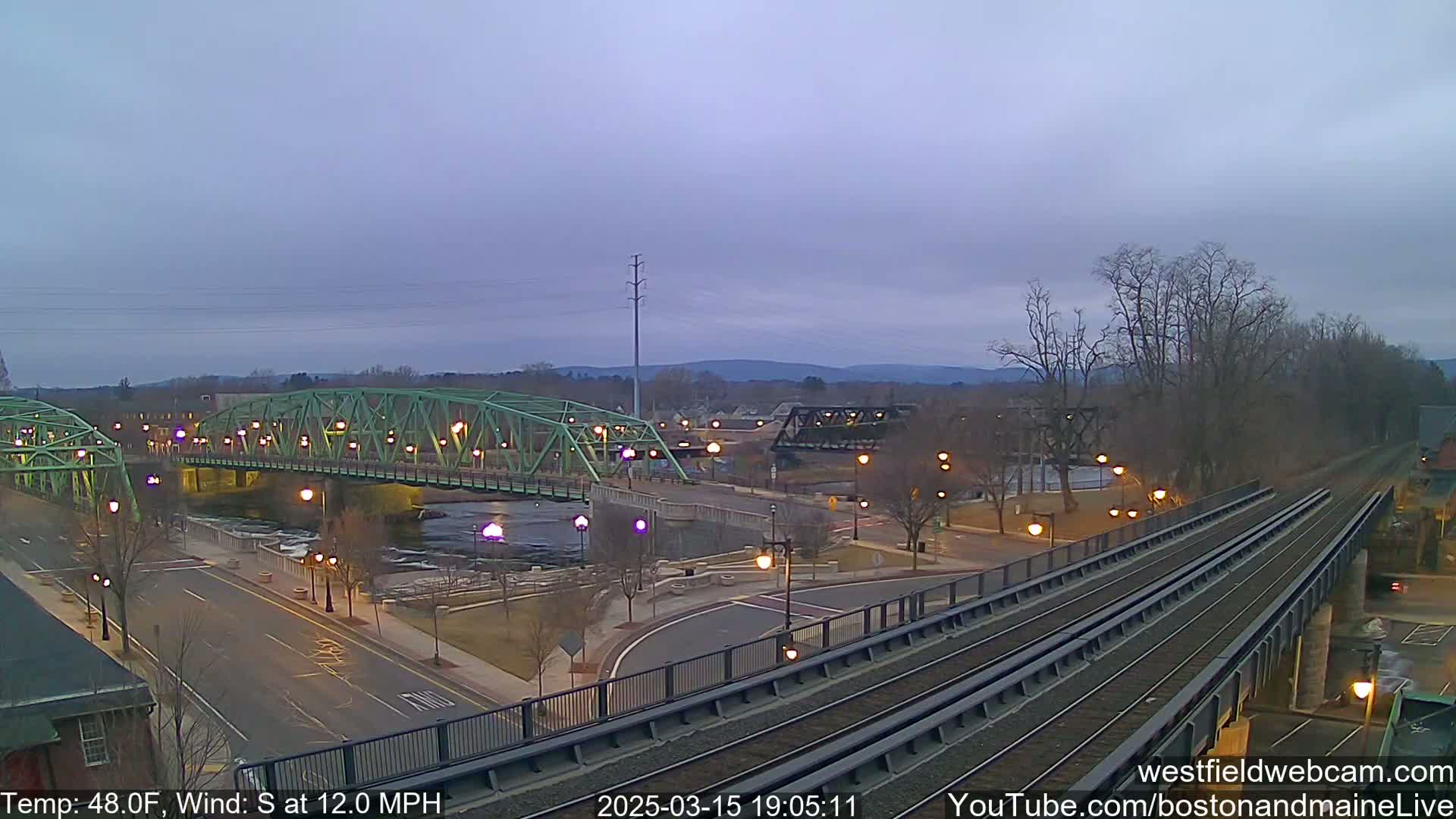 A green bridge spans a river beside a train trestle under a twilight sky with a south wind at 12 mph and a temperature of 48°F.