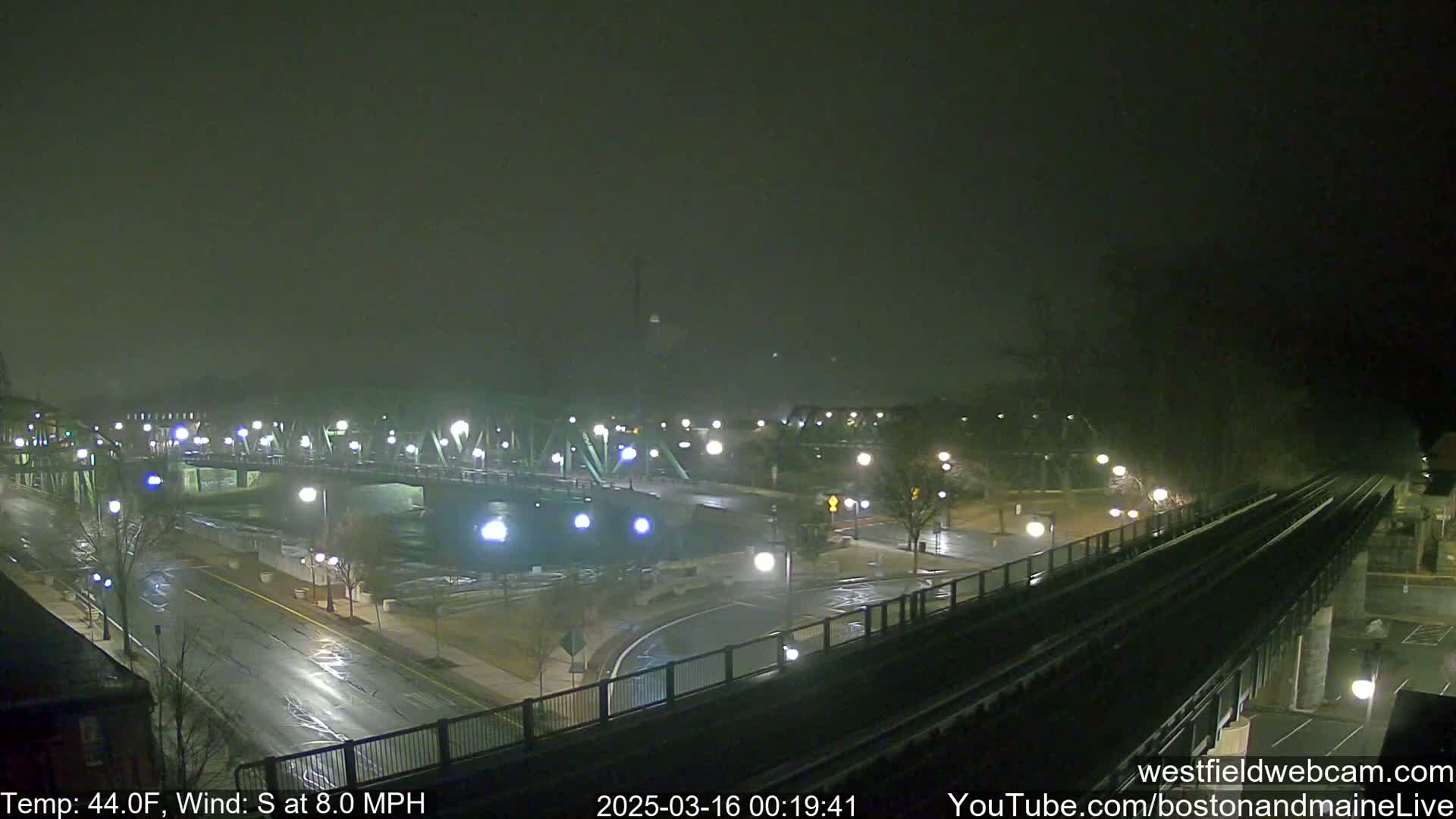 A nighttime view of a bridge and roadway illuminated by streetlights under foggy conditions, with a train track visible in the foreground.