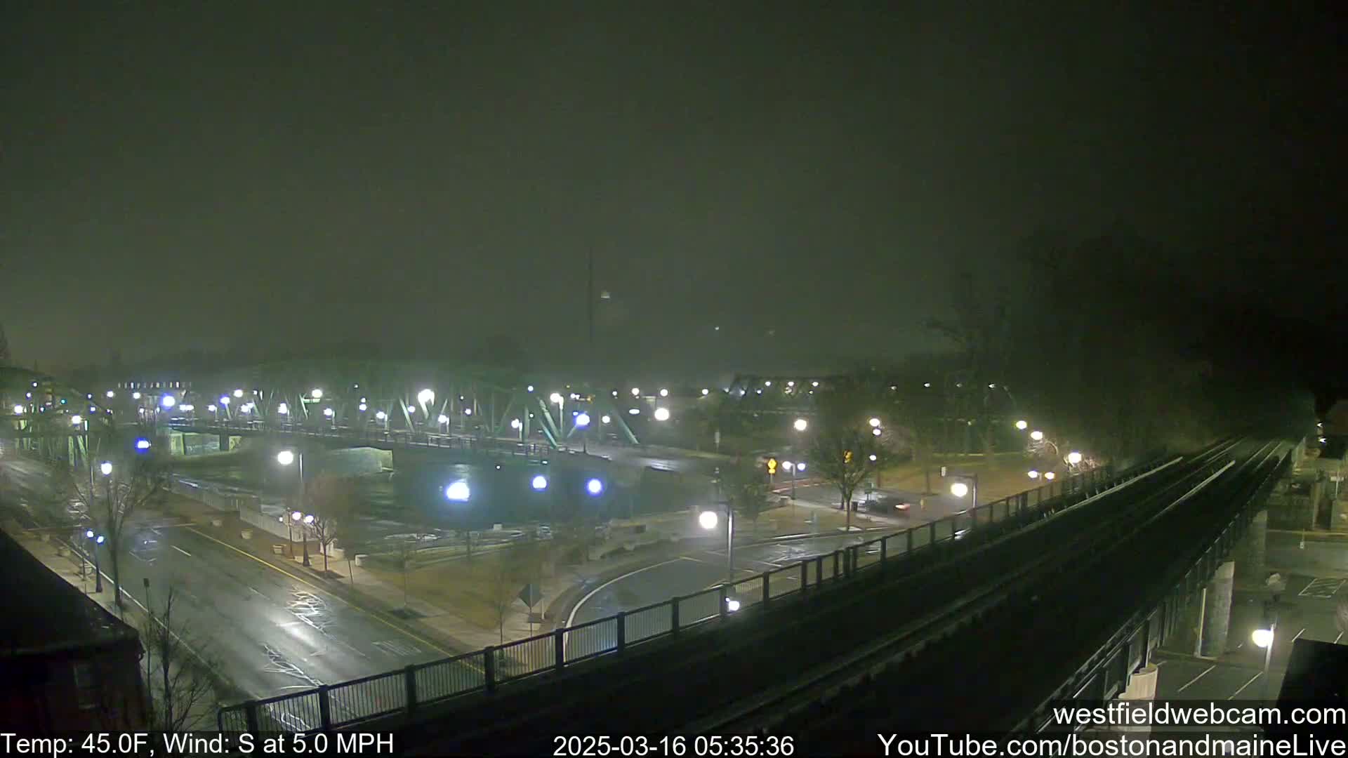 A nighttime, foggy view of a bridge and intersecting roadways with sparse traffic, under a sky obscured by fog or mist.