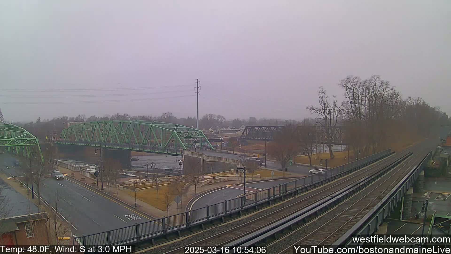 A view of two bridges over a river, with a railway line and roadway intersecting in the foreground, under an overcast sky.