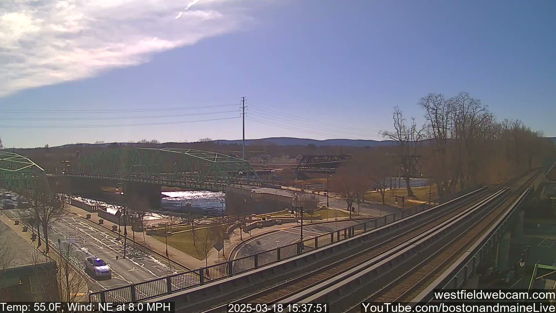 A view of a river with two bridges, one green and one dark-colored, under a partly sunny sky with a northeast wind at 8.0 mph, shows a roadway with a car and train tracks in the foreground.