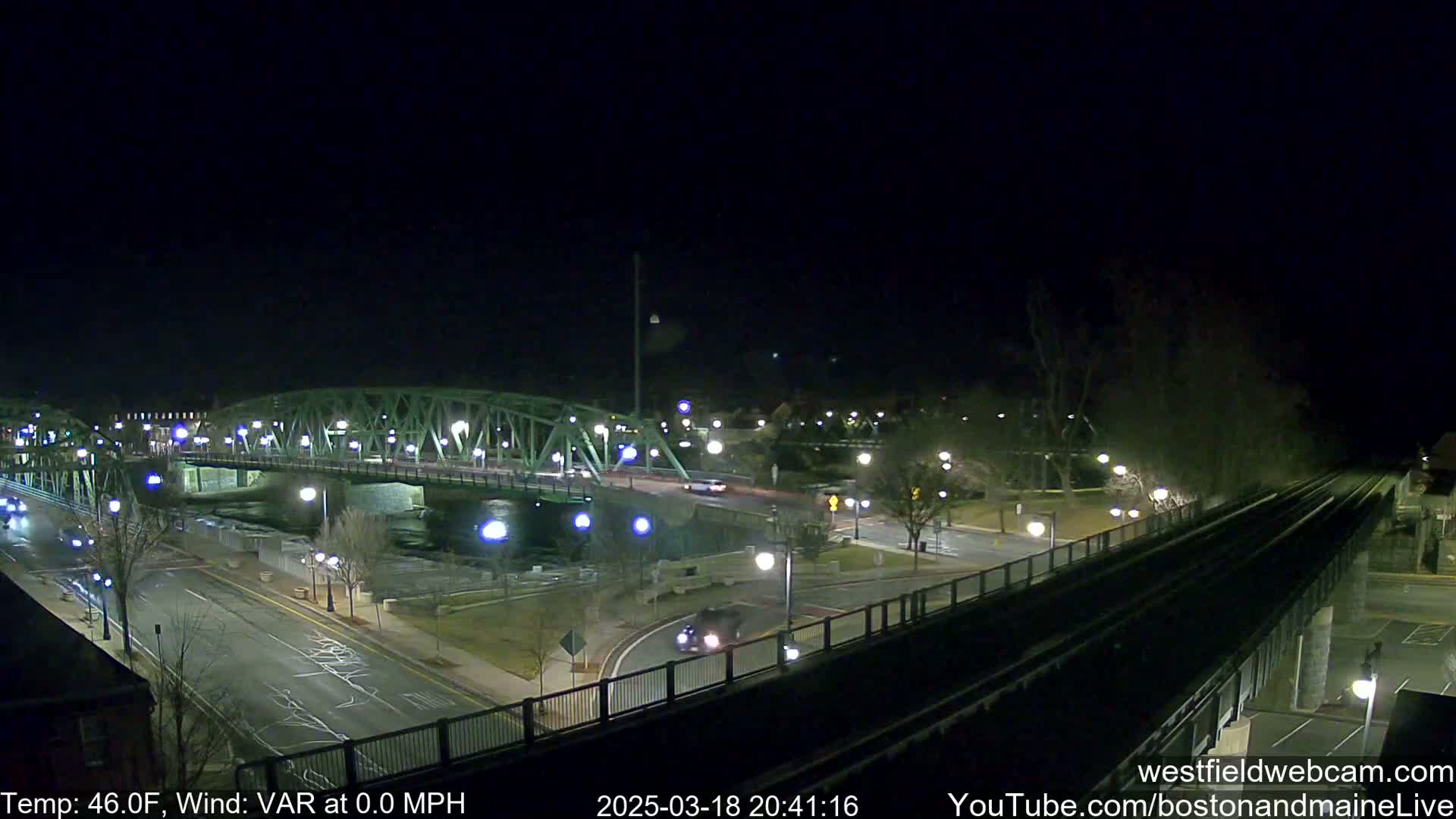 A nighttime view of a green metal bridge over a river, with cars on a road and train tracks visible, under a clear sky.