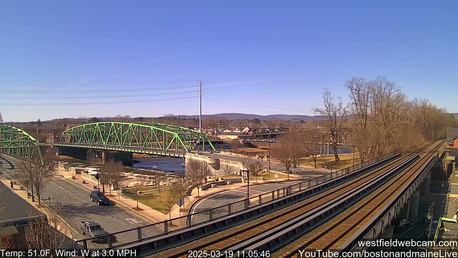 A green bridge spans a river under a clear blue sky, with train tracks and a road visible in the foreground, and bare trees lining the riverbank.