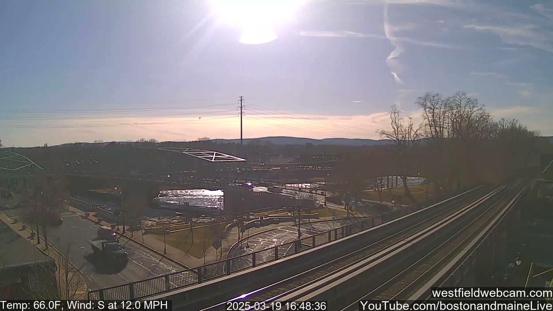 A sunny day with a south wind at 12 mph shows a view of a bridge over a river, a park, a roadway with vehicles, and train tracks in the foreground.