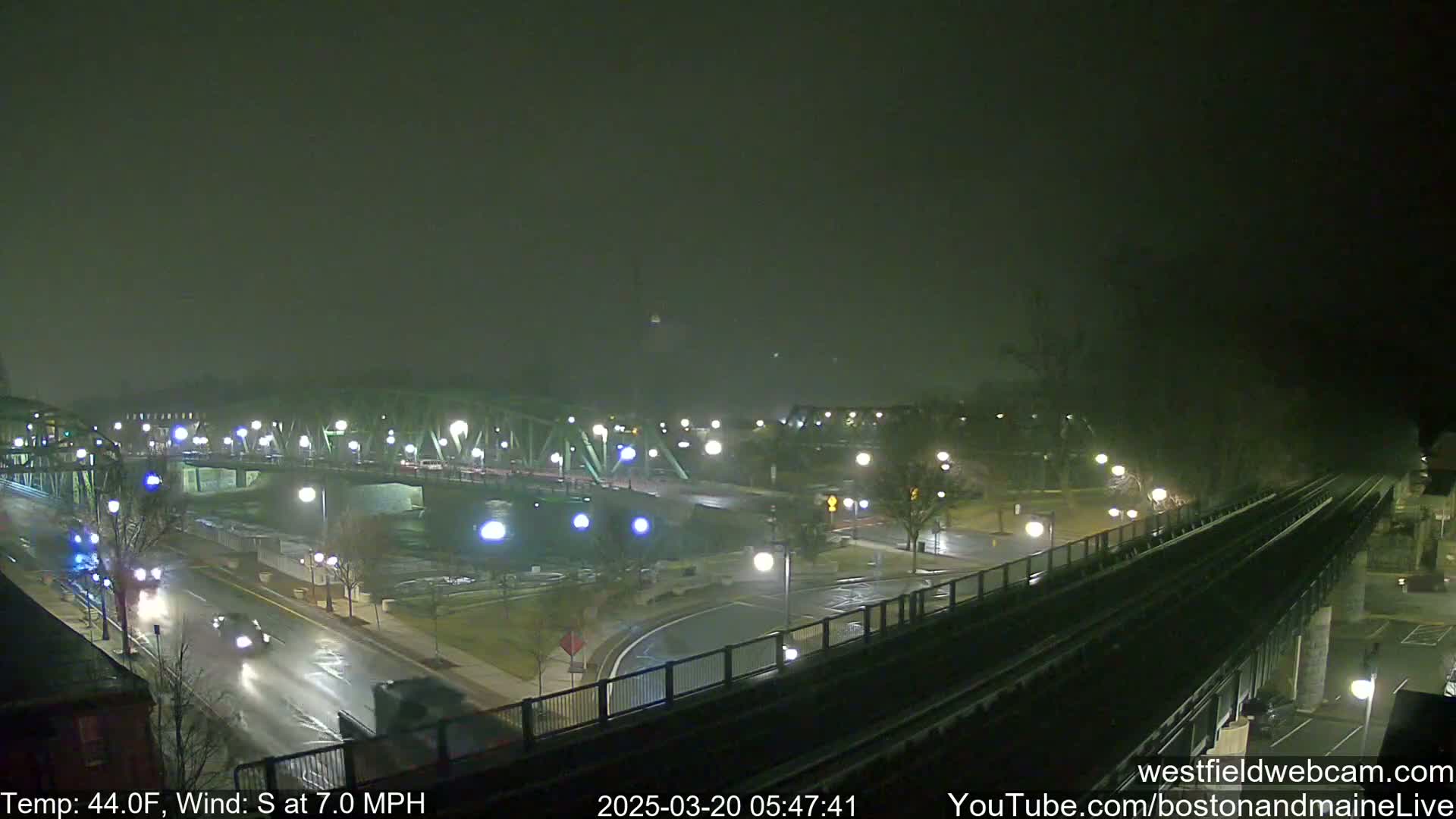 A nighttime view of a bridge and roadway with sparse traffic under foggy conditions, showing a train track elevated above the road.