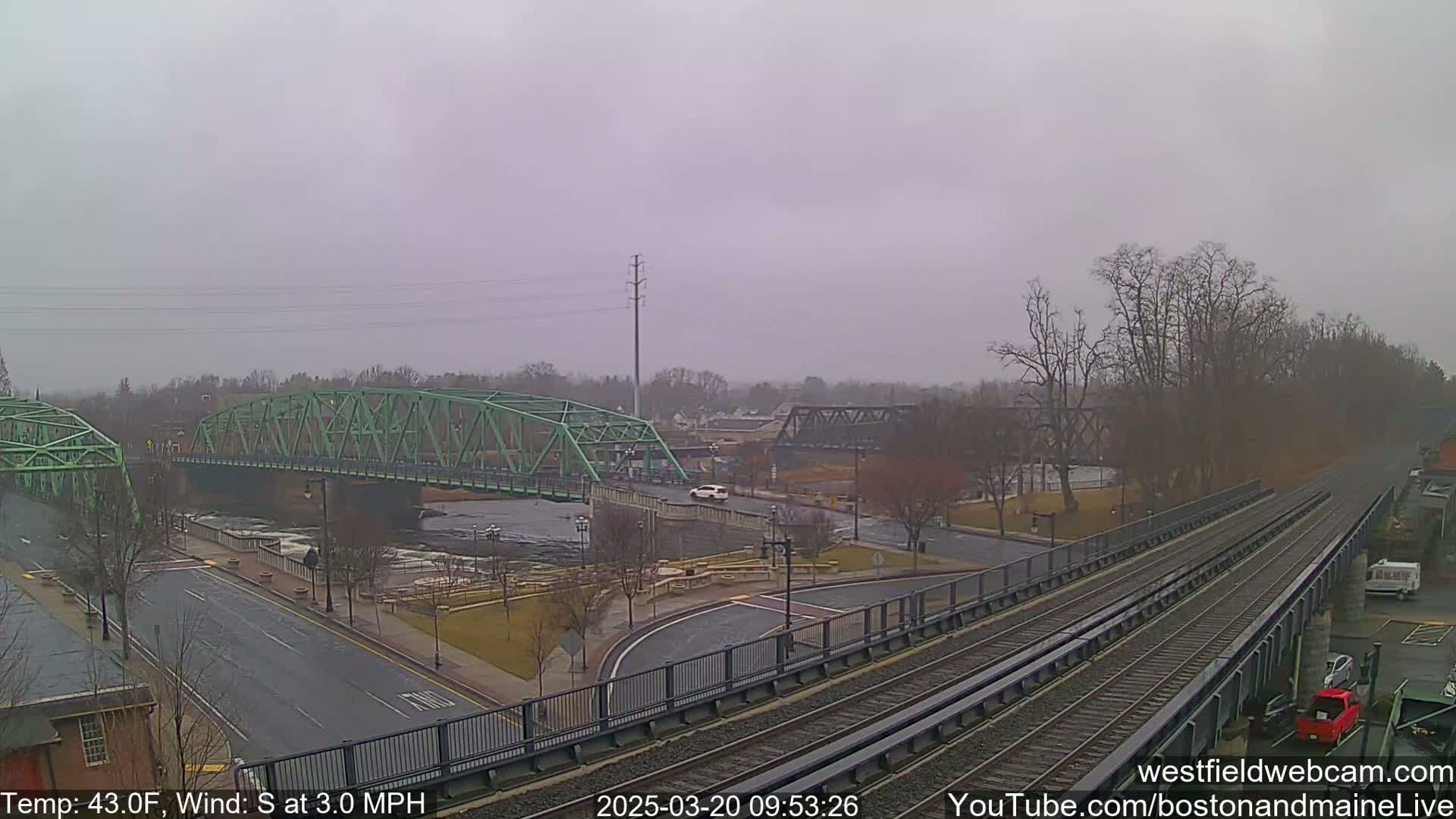 A green bridge spans a river under an overcast sky, with train tracks and a road visible in the foreground.