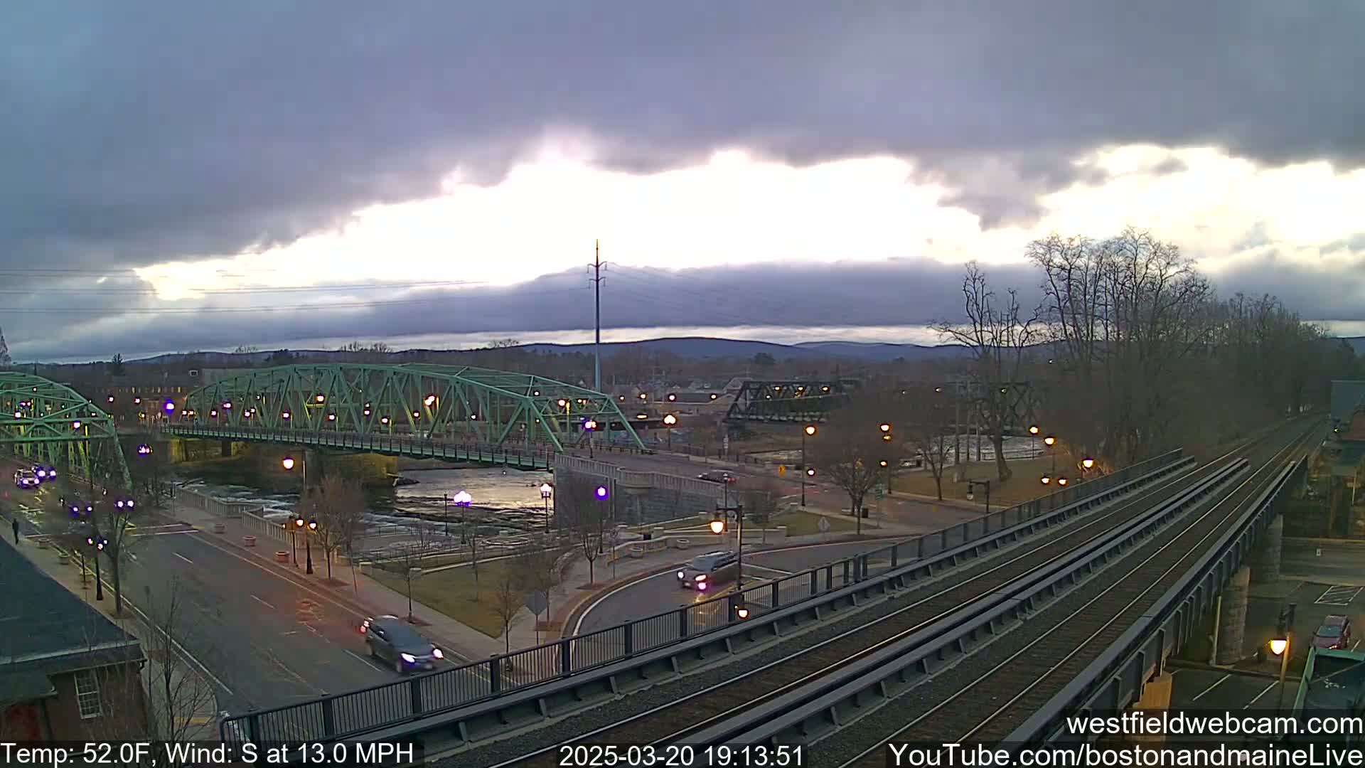 A view of a river, a green bridge, and train tracks under a partly cloudy sky.