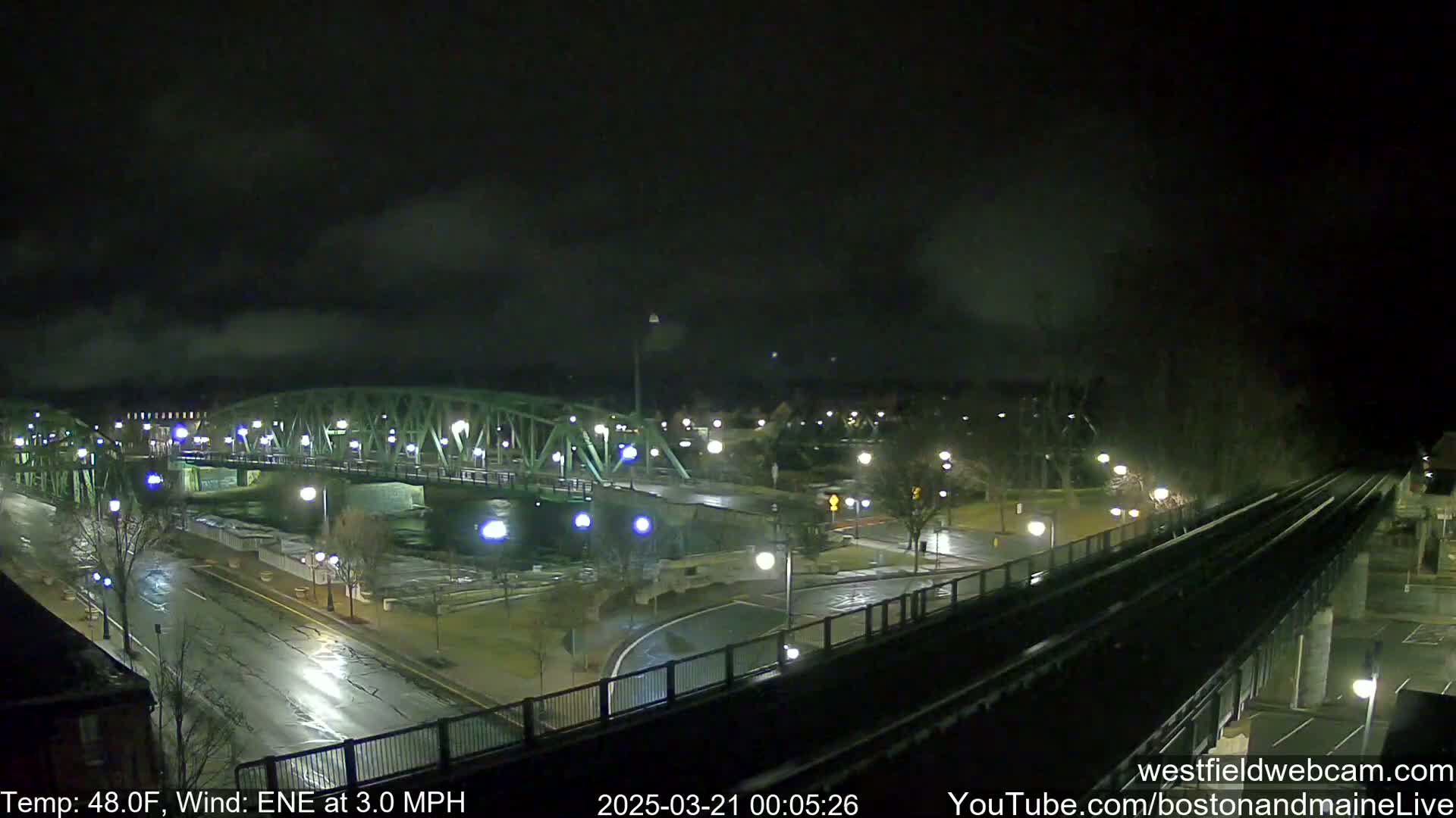 A green metal bridge spans a dark river at night under a cloudy sky, with wet roads and train tracks visible below.