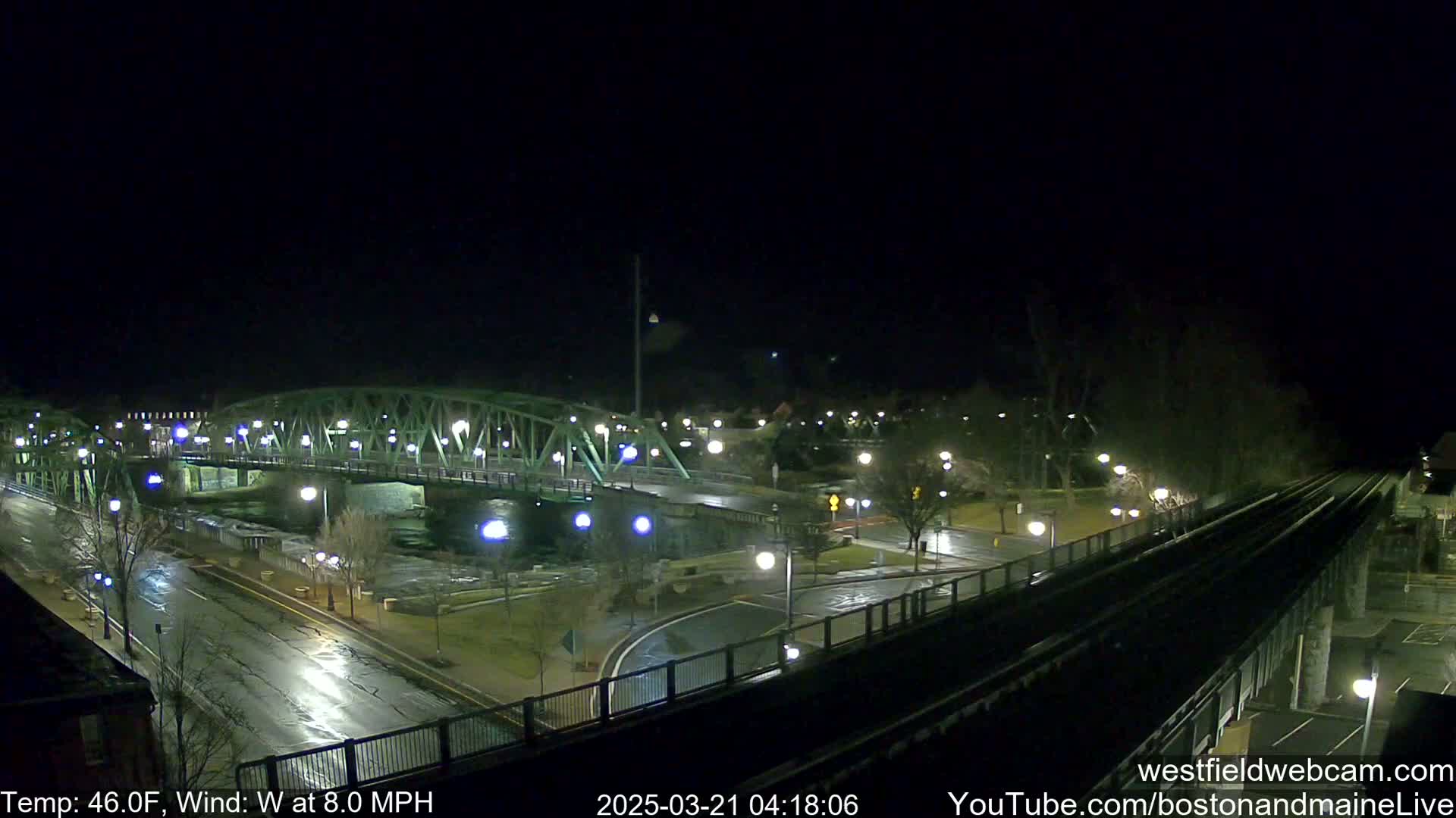 A nighttime view of a bridge over a river, with wet roads and a train track in the foreground, under a dark sky with a westerly wind.