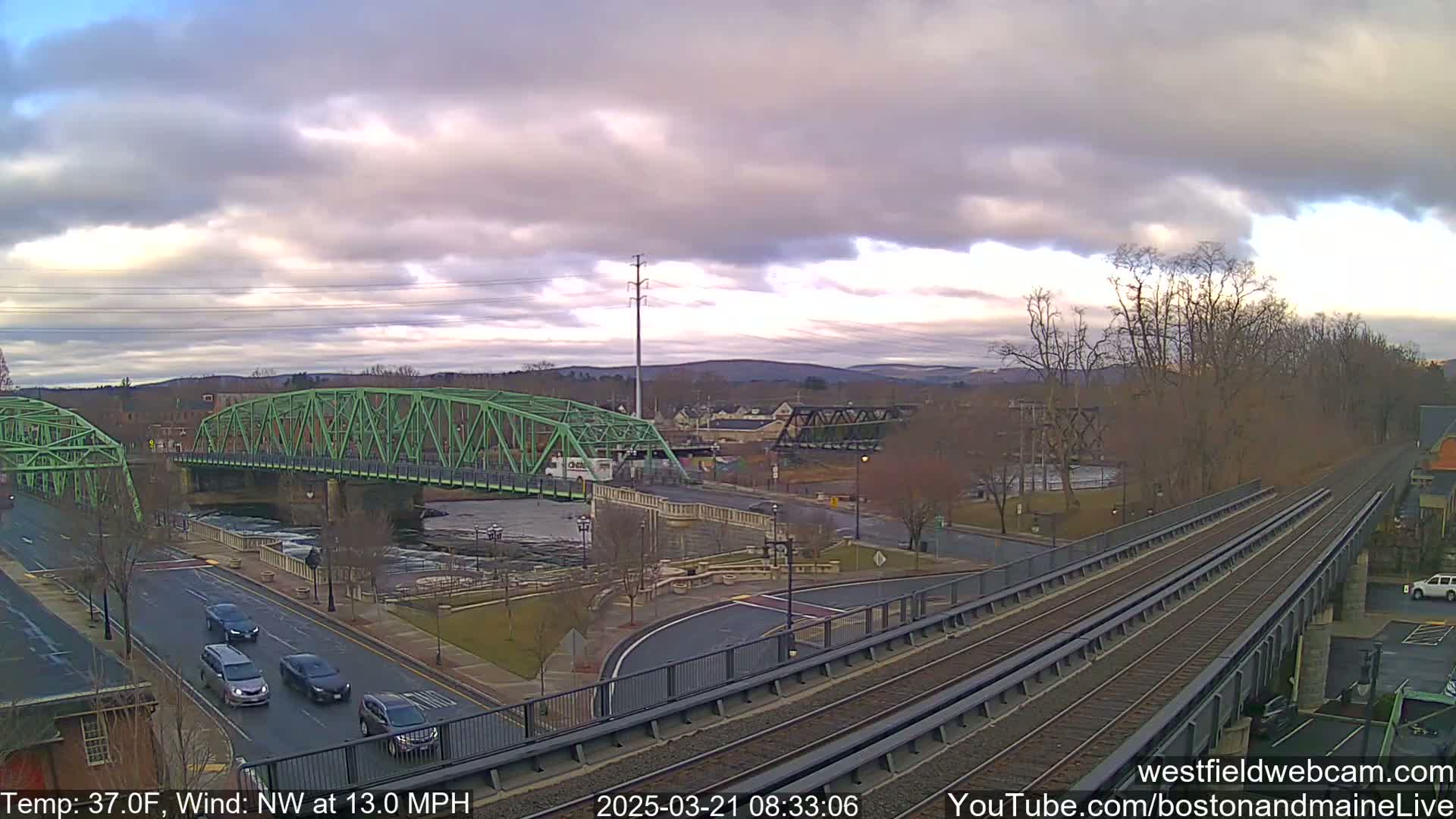 A view of a town under a cloudy sky, showing a green bridge over a river, a road with cars, and train tracks leading to a higher elevation.