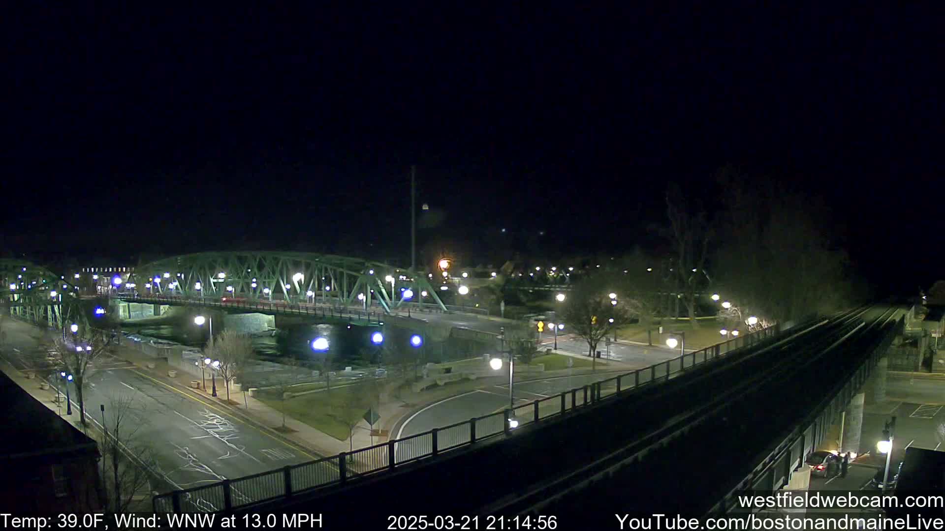 A nighttime view of a bridge over a river, with a train track elevated alongside a road, under a clear, dark sky and a 39°F temperature with a 13 mph wind from the west-northwest.