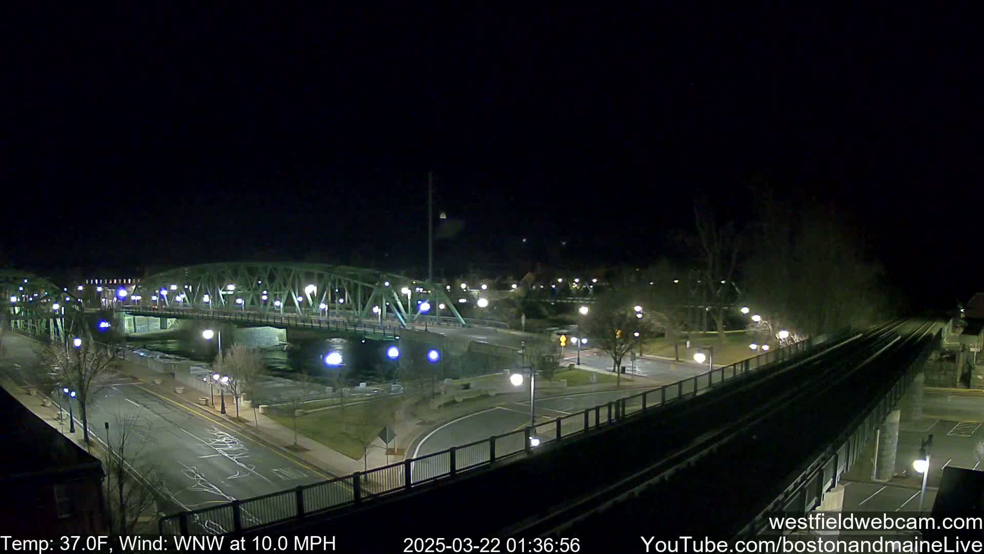 A nighttime view of a bridge over a river, with a street and train tracks visible, under a clear, dark sky with a temperature of 37°F and a west-northwest wind at 10 mph.