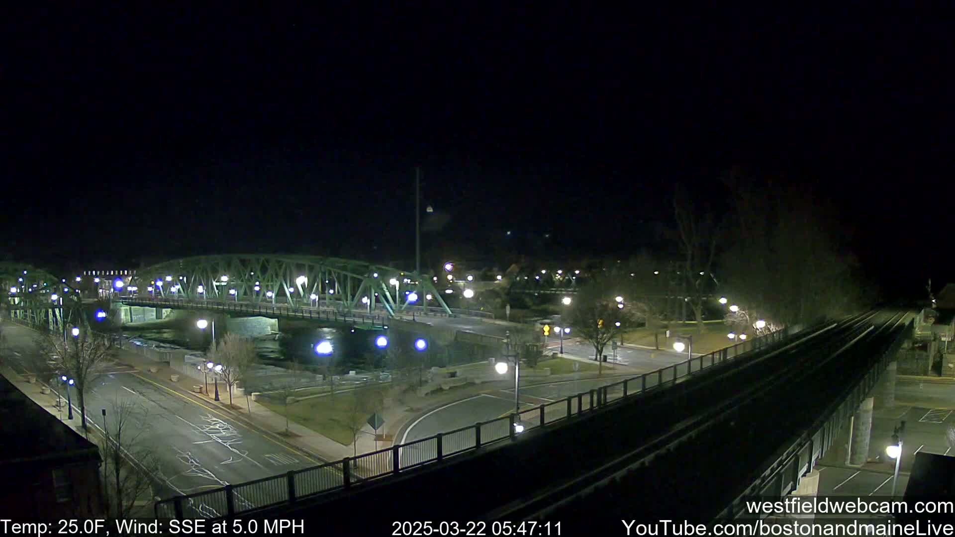 A nighttime view of a bridge over a river, with a road and train tracks visible, under a clear, dark sky with a temperature of 25 degrees Fahrenheit and a southeasterly wind of 5 mph.
