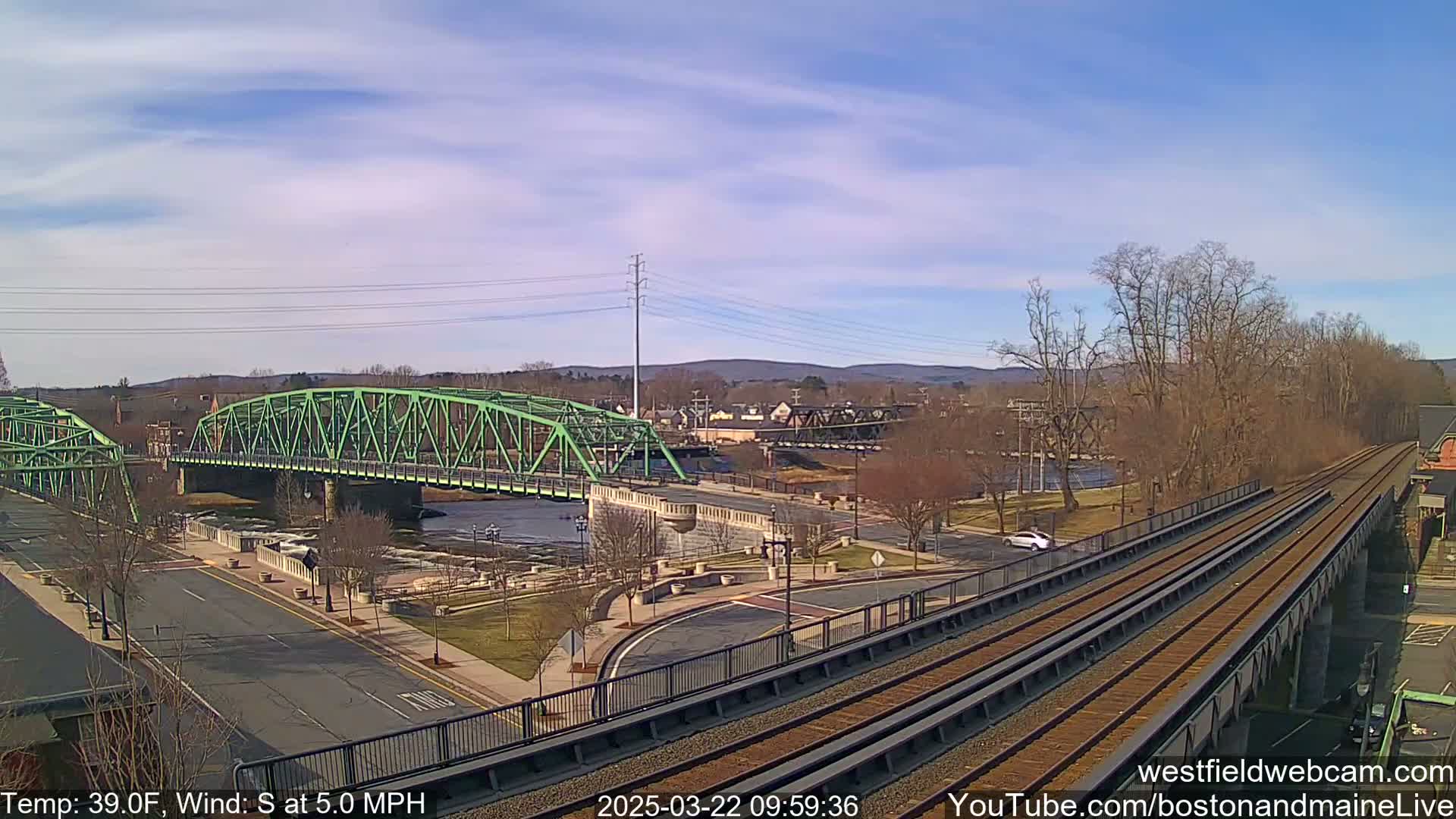 A green bridge spans a river in a town under a partly cloudy sky, with train tracks running alongside a road in the foreground.
