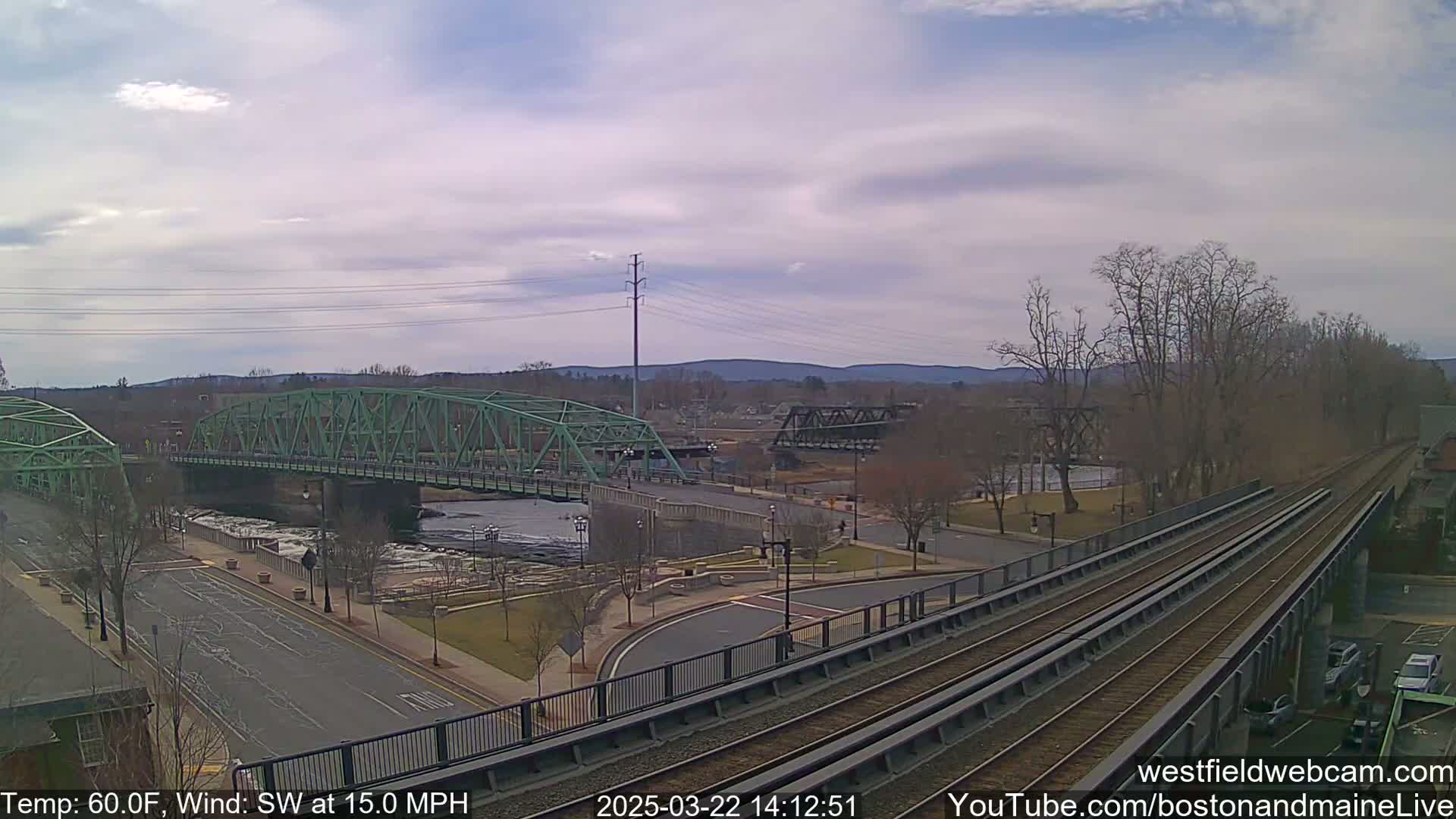 A view of a river with two bridges, one green and one dark metal, under a partly cloudy sky, with a train track in the foreground and a 15 mph southwest wind.