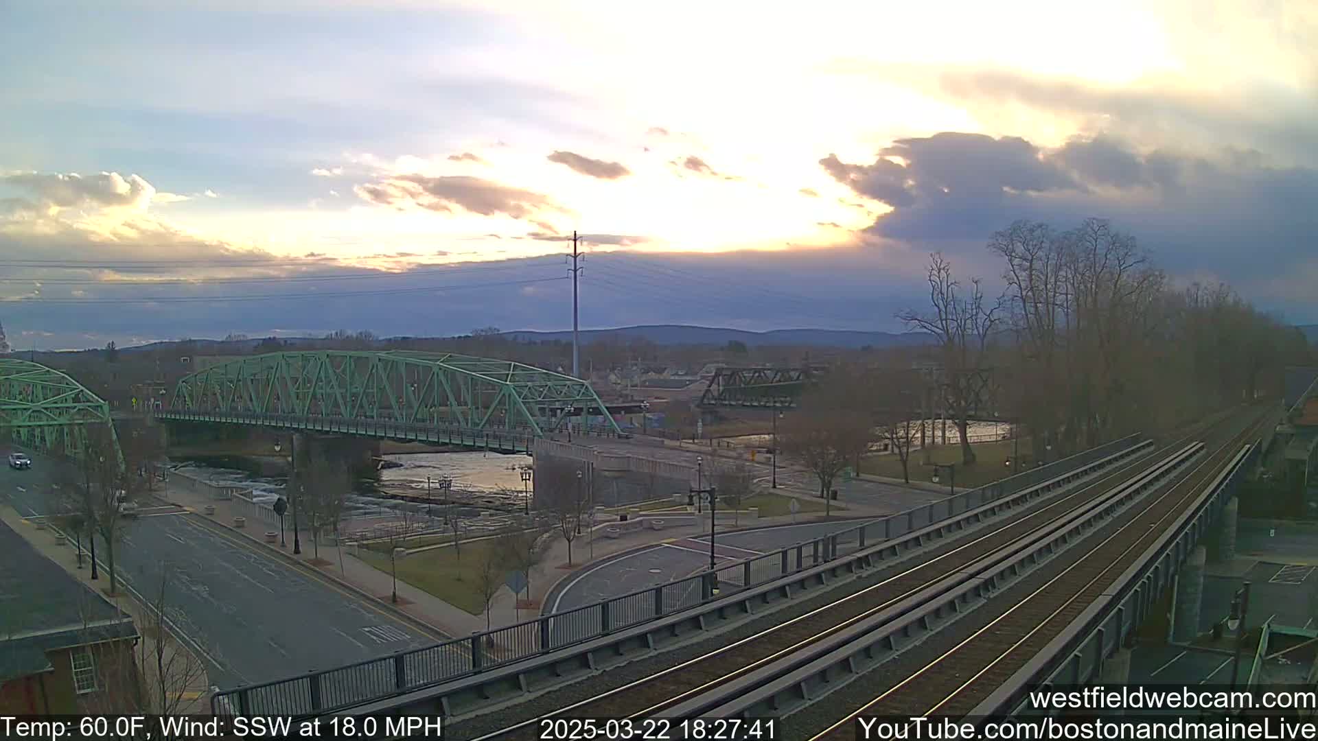 A green bridge spans a river under a partly cloudy sunset sky, with train tracks visible in the foreground and a road leading to the bridge.