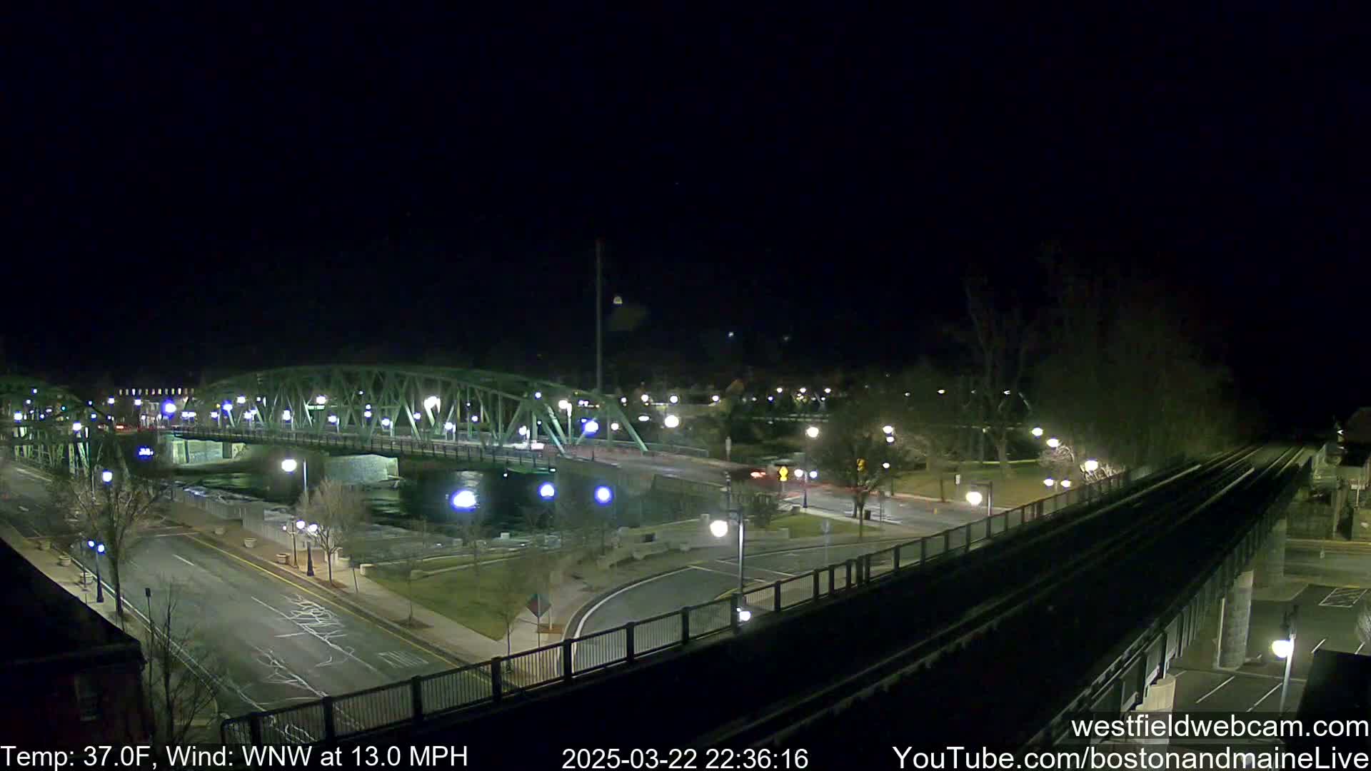 A nighttime view of a bridge over a river, with a road and train tracks visible, under a clear, dark sky with a temperature of 37 degrees Fahrenheit and a wind from the west-northwest at 13 miles per hour.