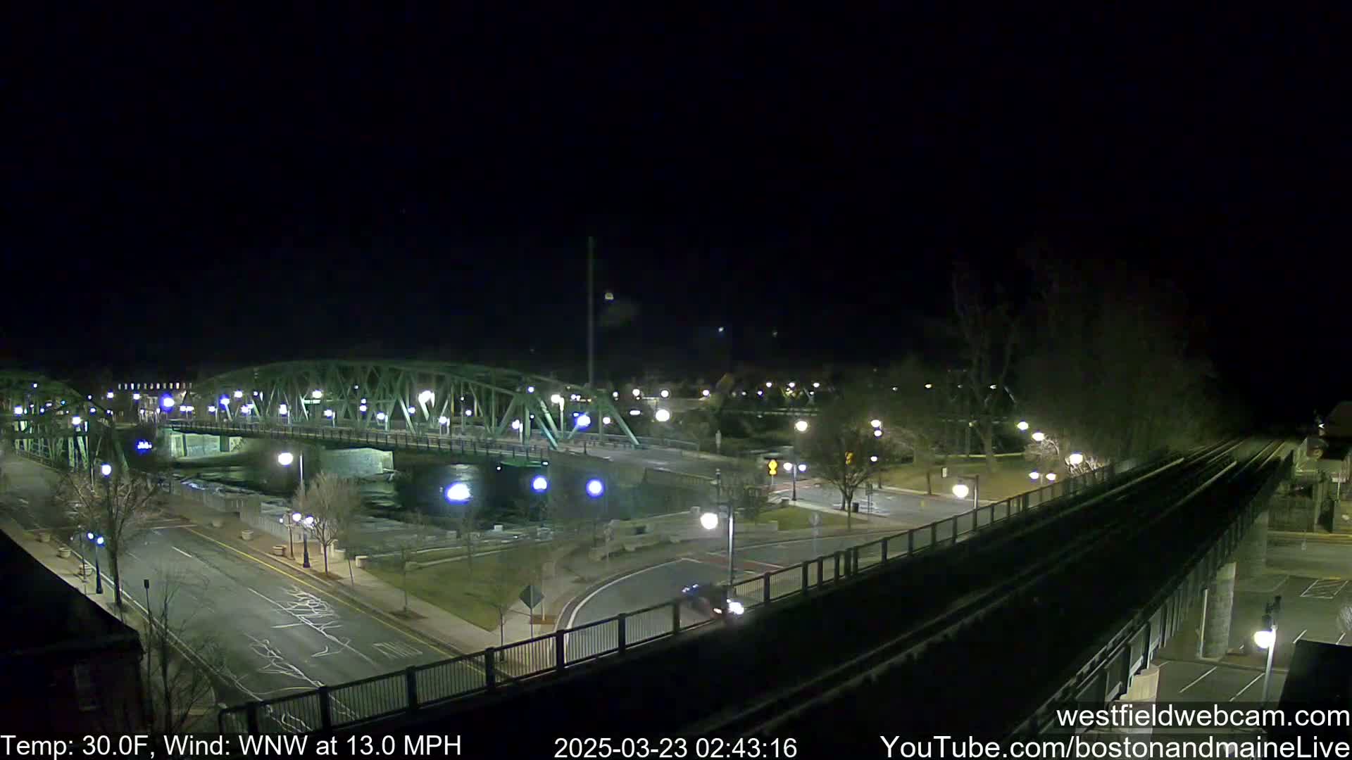 A nighttime view of a lit-up bridge over a river, with a road and train tracks below, under a clear, dark sky with a west-northwest wind at 13 miles per hour and a temperature of 30 degrees Fahrenheit.