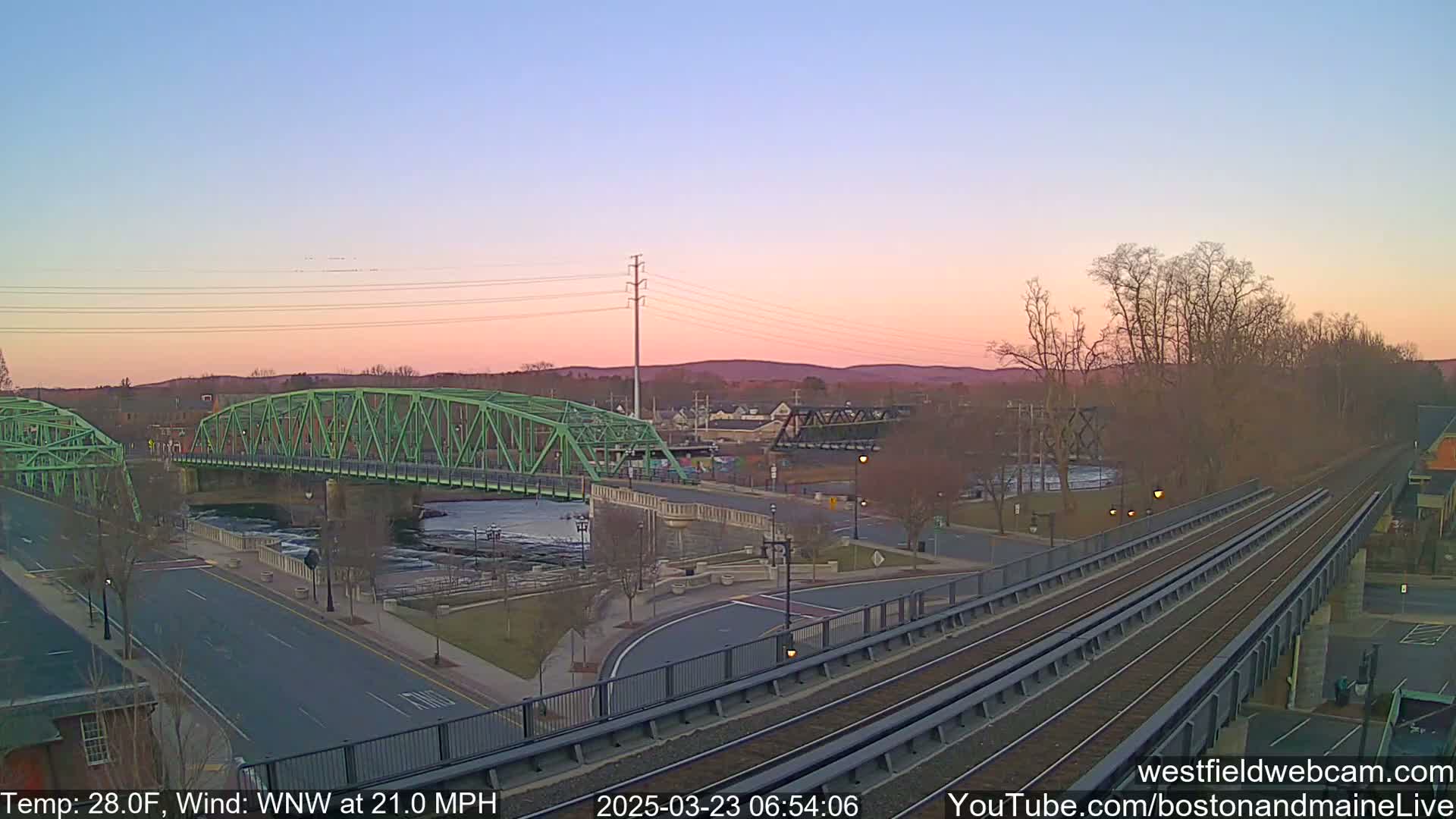 A pastel sunrise illuminates a river scene with two bridges, one green and one dark metal, alongside train tracks and a road.