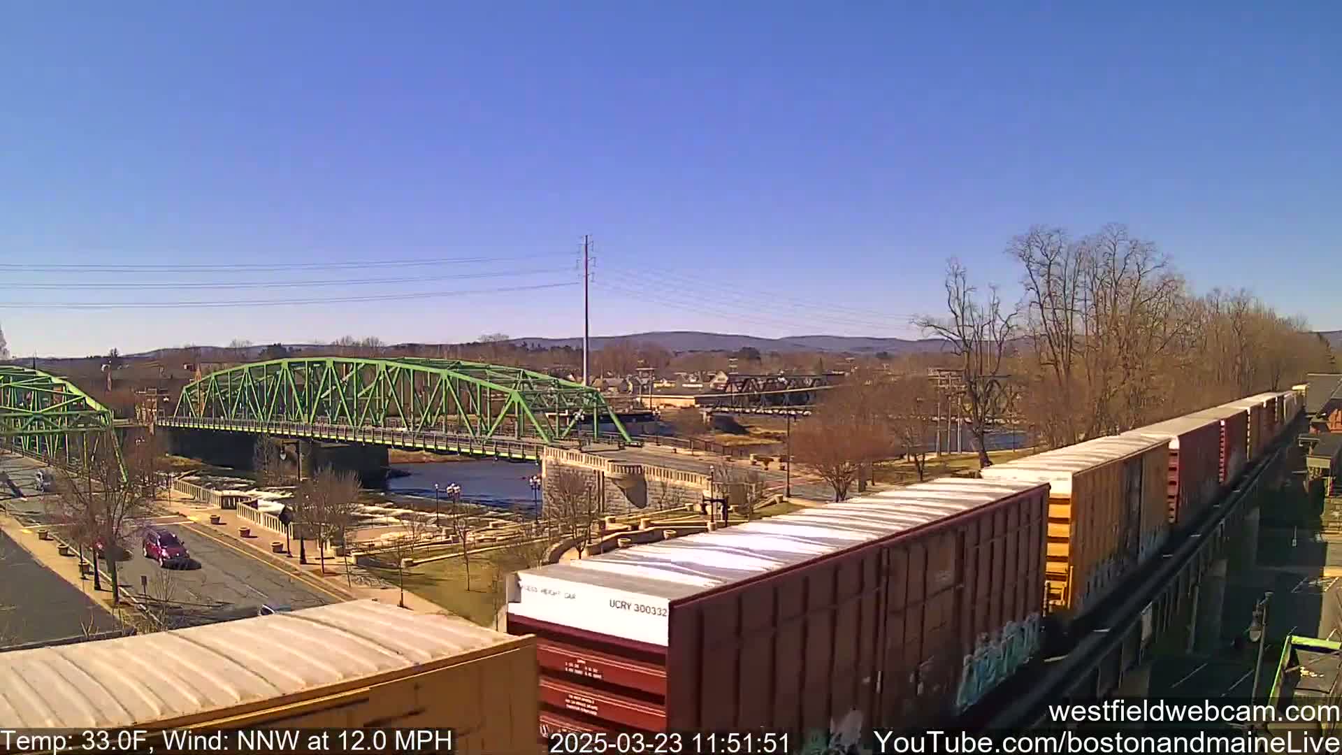 A green bridge spans a river, with a train passing in the foreground on a sunny day with a north-northwest wind at 12 miles per hour.
