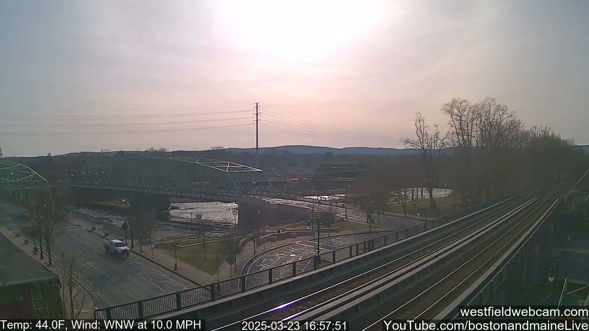 A partly cloudy, breezy day reveals a river scene with two bridges, a roadway, and train tracks, all under a pale sky.