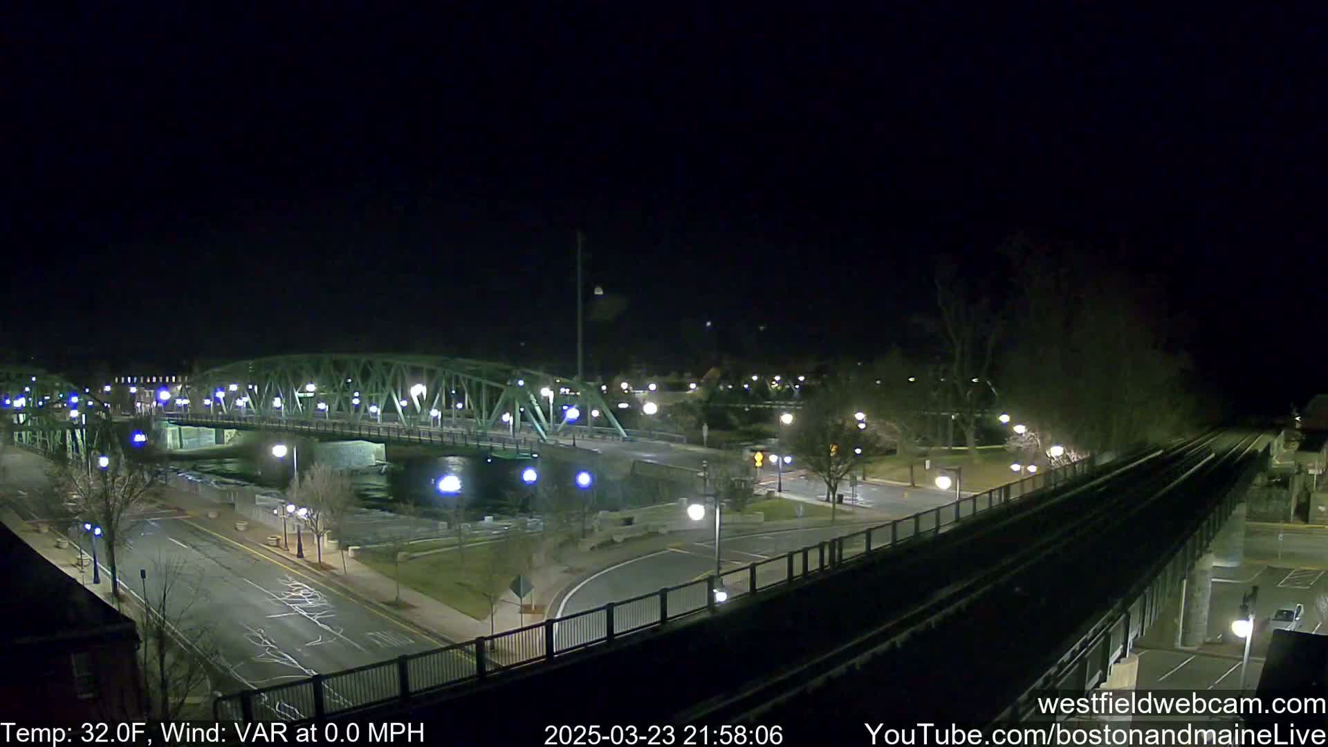 A nighttime view of a bridge over a river, a road, and train tracks, with a temperature of 32 degrees Fahrenheit and no wind.