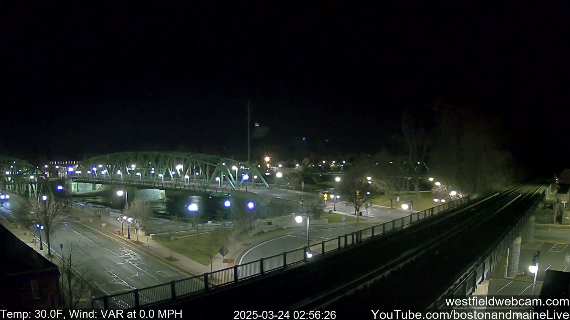 A nighttime view of a bridge over a river, with a road and train tracks below, under clear skies and a temperature of 30 degrees Fahrenheit.