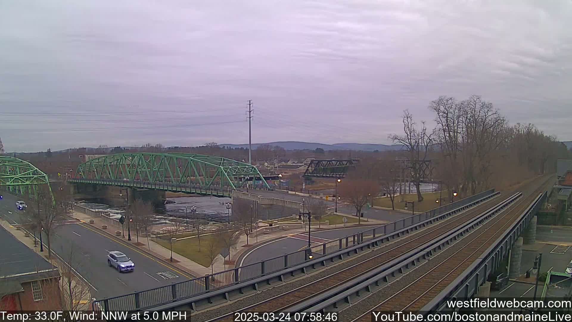 A view of two bridges over a river, with train tracks in the foreground, under a cloudy sky.