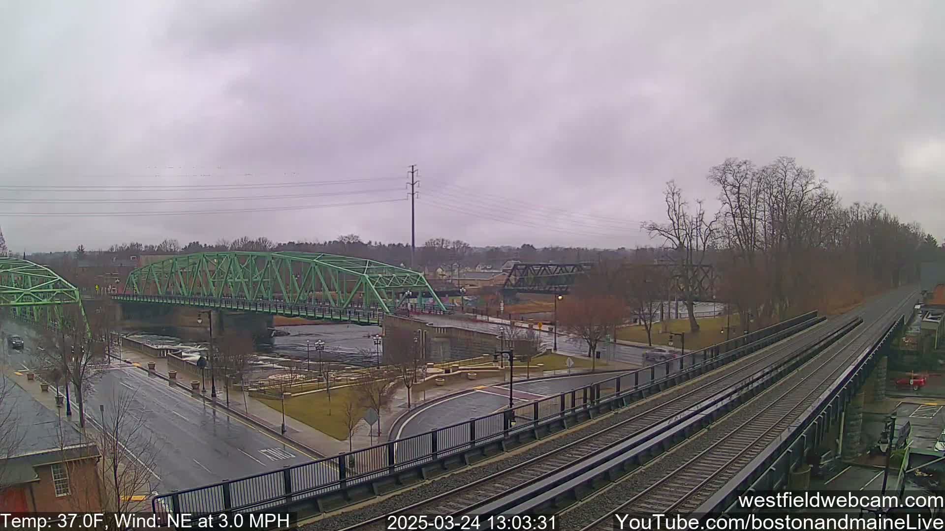 A view of a river with two bridges, one green and one dark metal, under an overcast sky, with train tracks in the foreground and a road running alongside the river.
