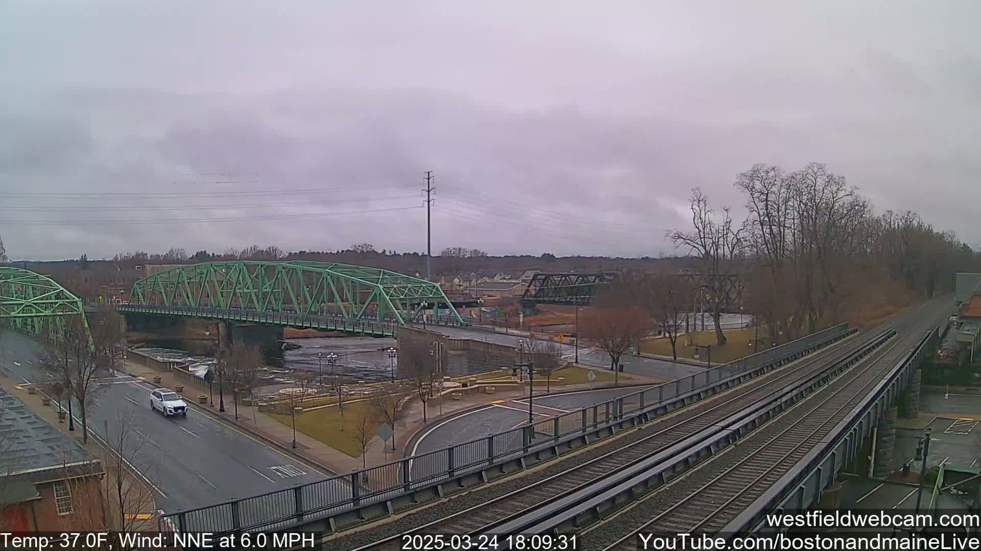 A view of a river with two bridges, one green and one black, under an overcast sky with a light wind.