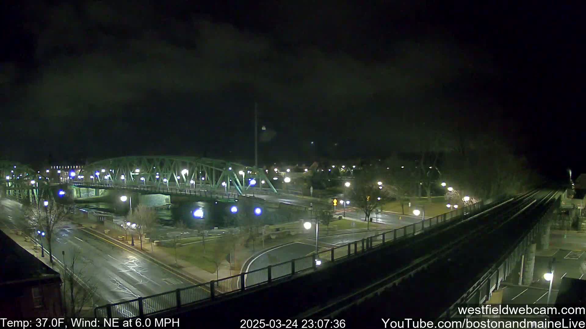 A nighttime view of a bridge over a river, with a roadway and train tracks visible under a dark, cloudy sky with a northeast wind.