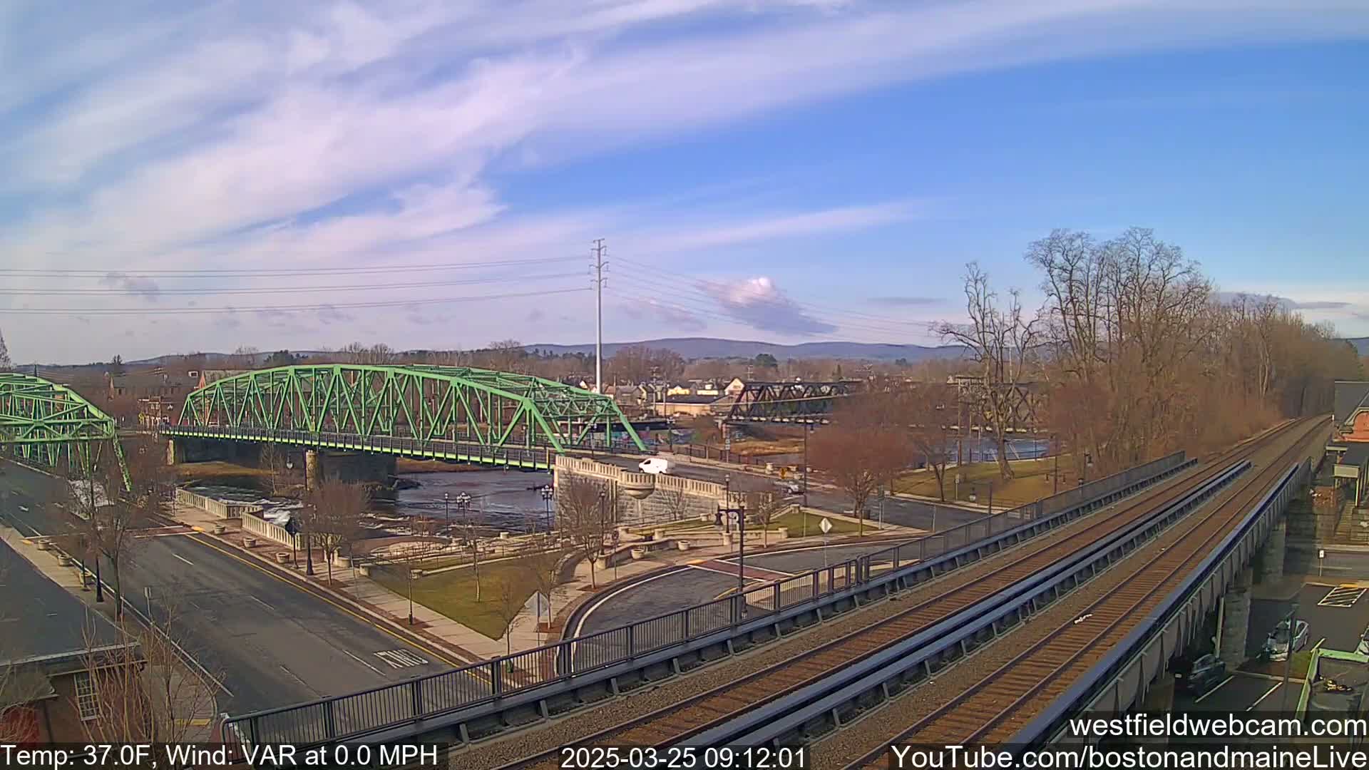 A partly cloudy day reveals a green metal bridge over a river, with train tracks and a town in the background.