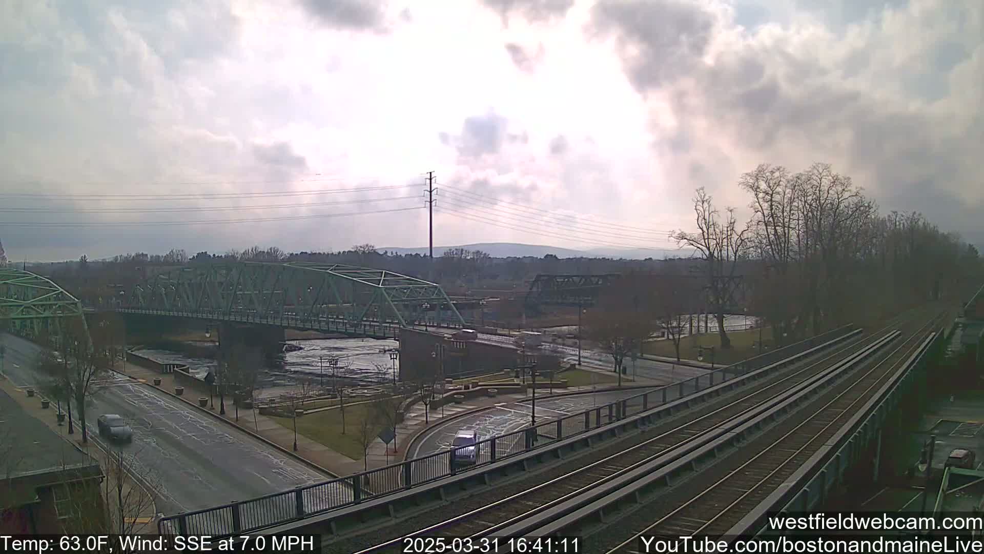 A partly cloudy day reveals a river scene with two bridges, a road, train tracks, and sparse early spring vegetation.