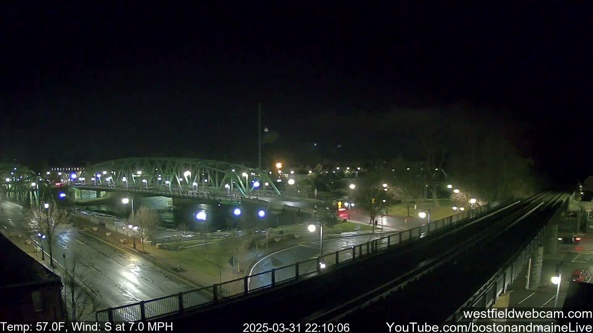 A nighttime view of a green bridge over a river, with wet streets and a train track visible, under a clear sky with a south wind.