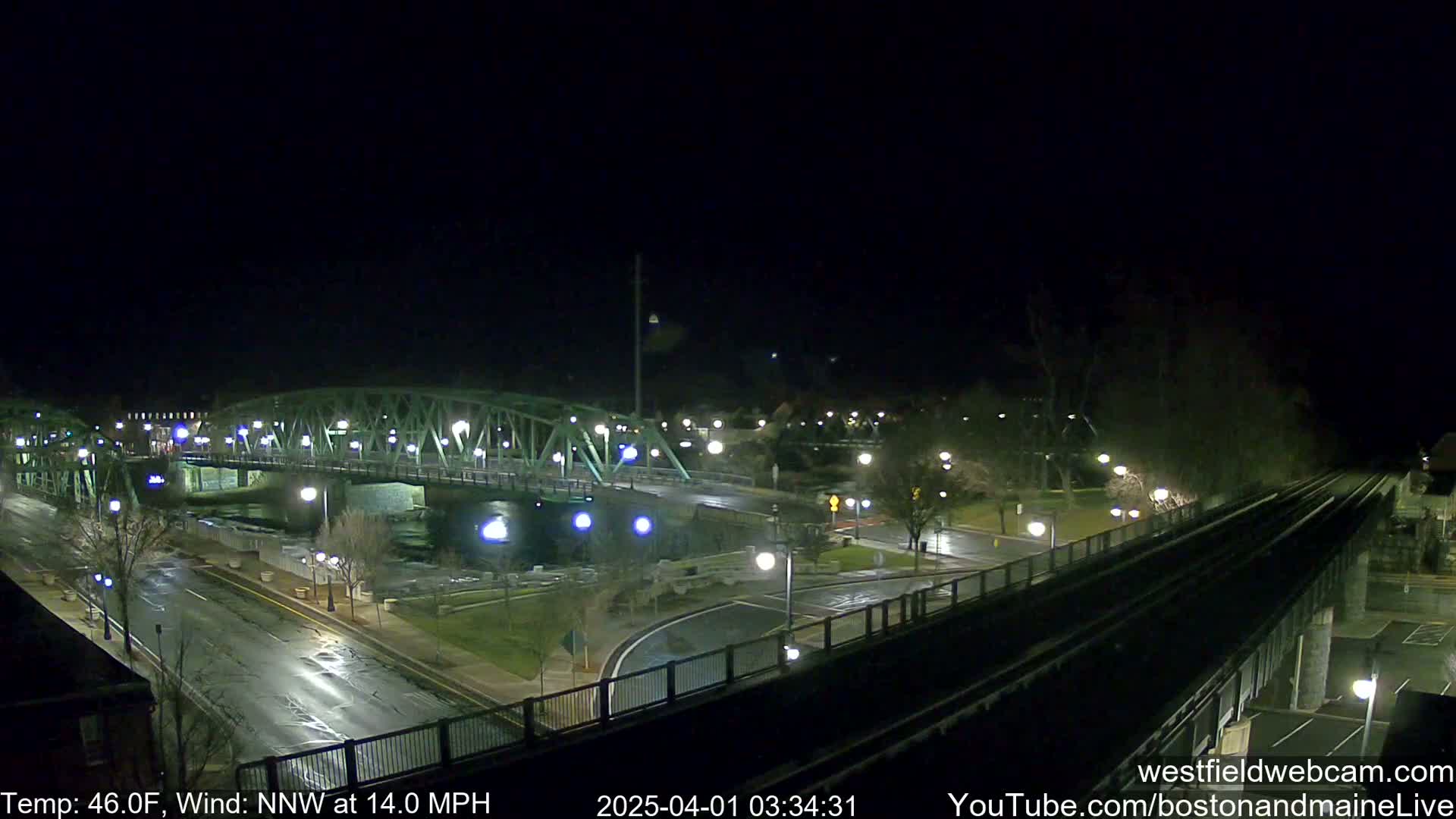 A nighttime view of a bridge over a river, with a road and train tracks visible, under a dark sky with a 46°F temperature and a 14 mph north-northwest wind.