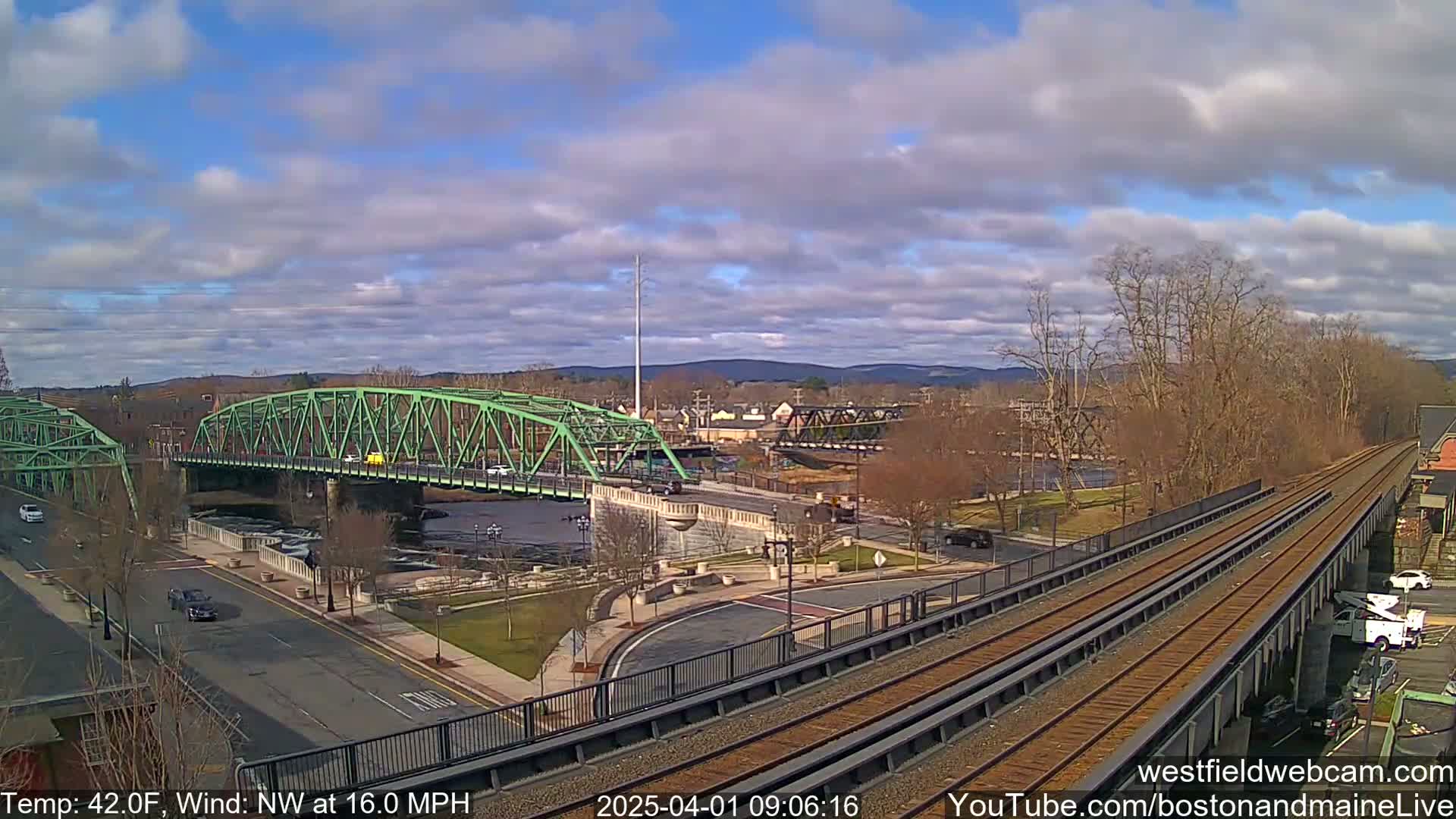 A green bridge spans a river under a partly cloudy sky, with train tracks visible in the foreground and a town in the background.