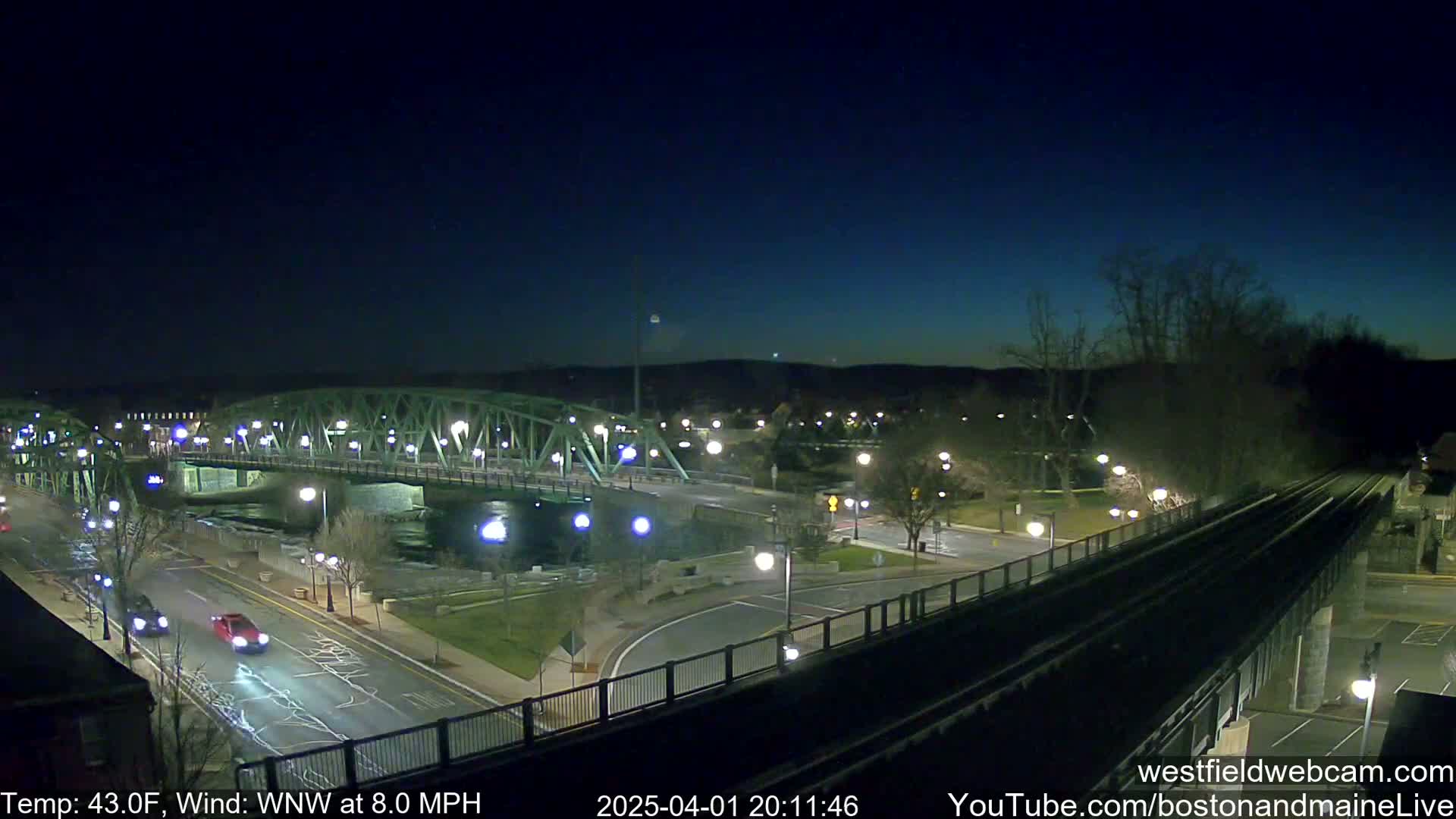 A nighttime view of a river with a green bridge and roadways, under a mostly clear sky with a temperature of 43 degrees Fahrenheit and a west-northwest wind at 8 miles per hour.