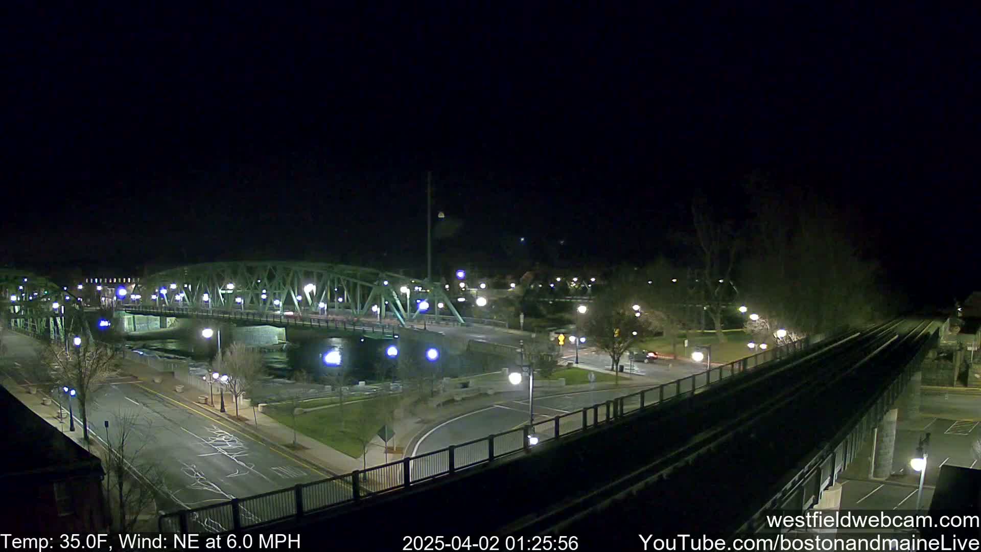 A nighttime view of a bridge over a river, with a road and train tracks intersecting, under a clear, dark sky and a north-easterly wind.