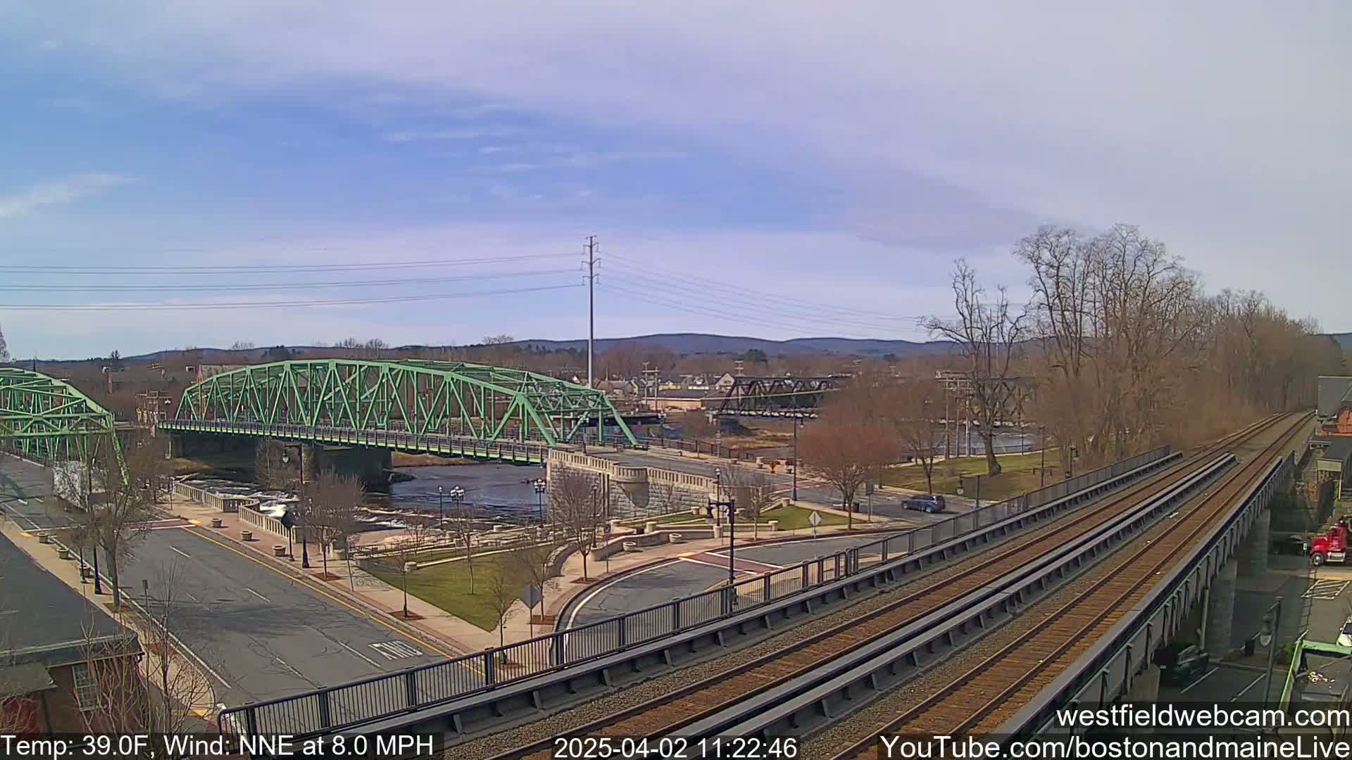 A green bridge spans a river, with train tracks and a road below, under a partly cloudy sky.