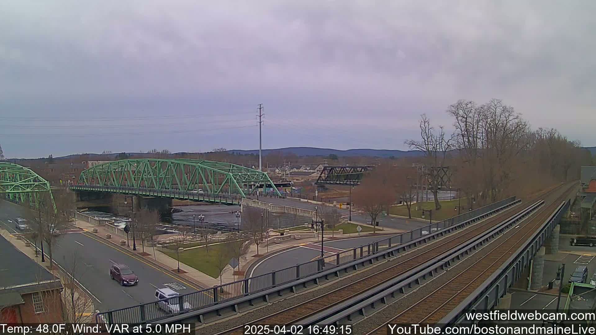 A view of a town with two bridges over a river, train tracks, and a road, under an overcast sky.
