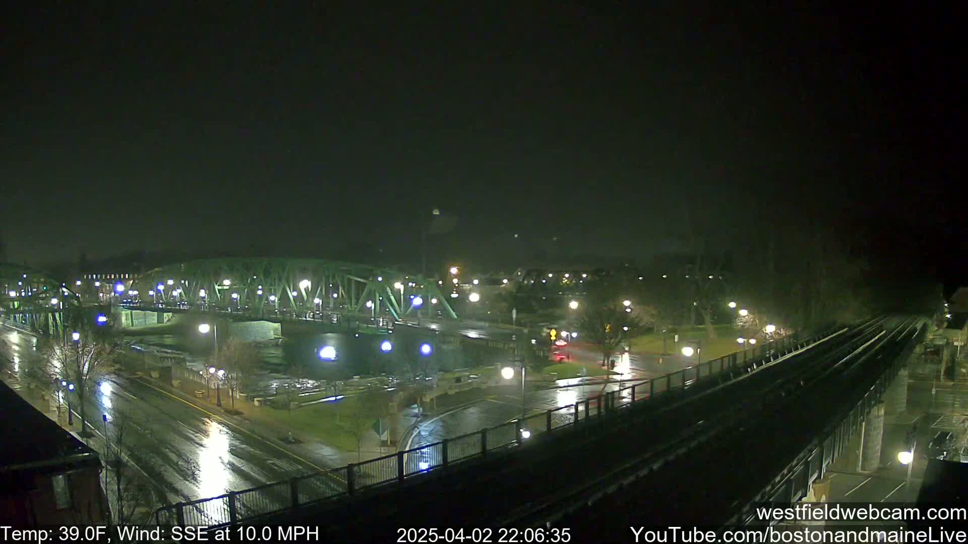 A rain-slicked nighttime scene shows a green bridge spanning a river, with wet streets and train tracks visible below, under a dark sky and a 10 mph wind from the south-southeast.