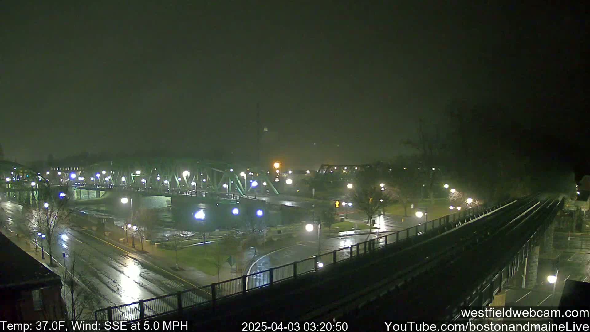A rain-slicked nighttime scene shows a bridge over a river, a road with streetlights, and a set of train tracks, all under a dark, overcast sky.