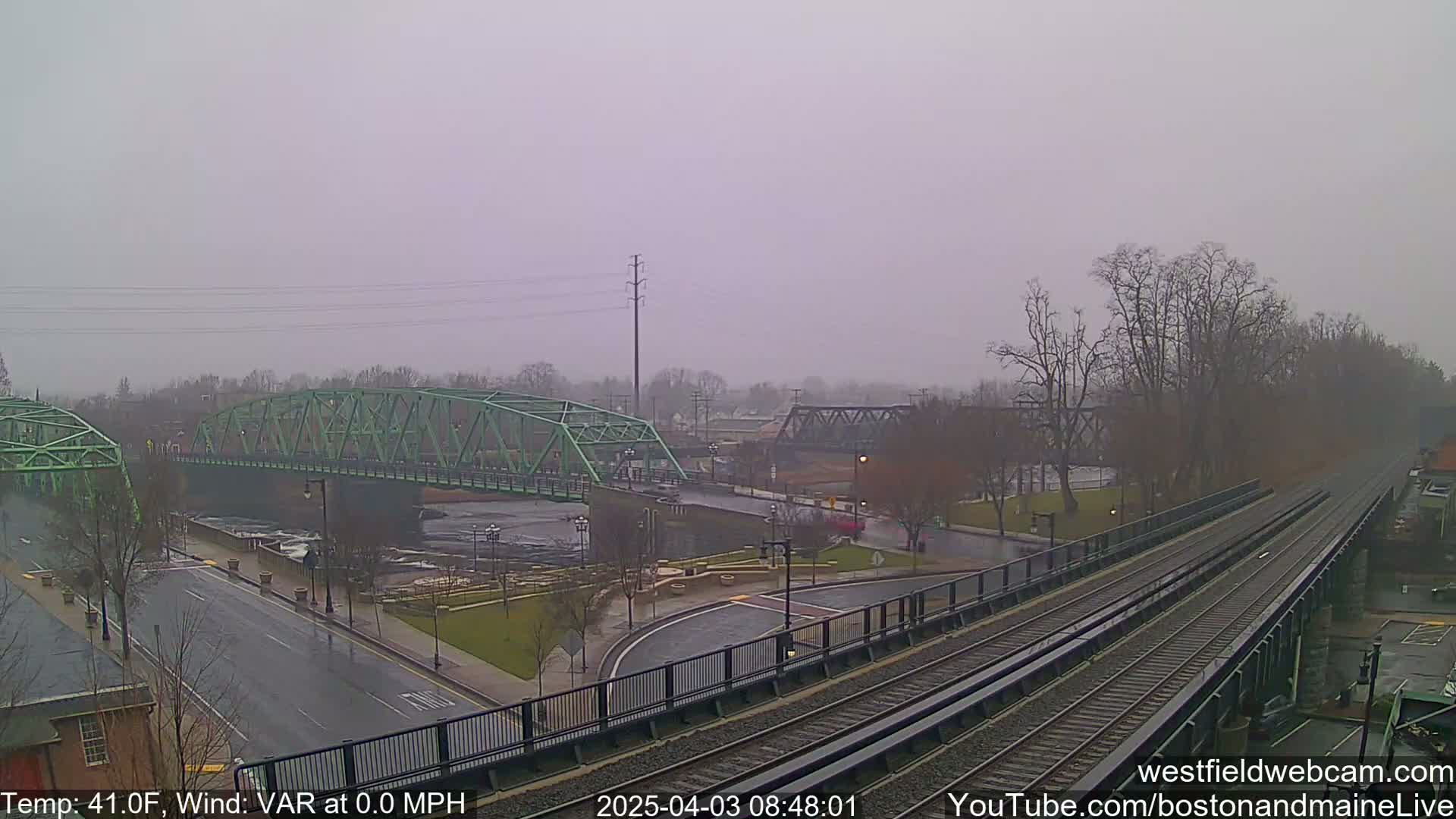 A view of a rain-soaked town with two bridges over a river, and train tracks in the foreground.