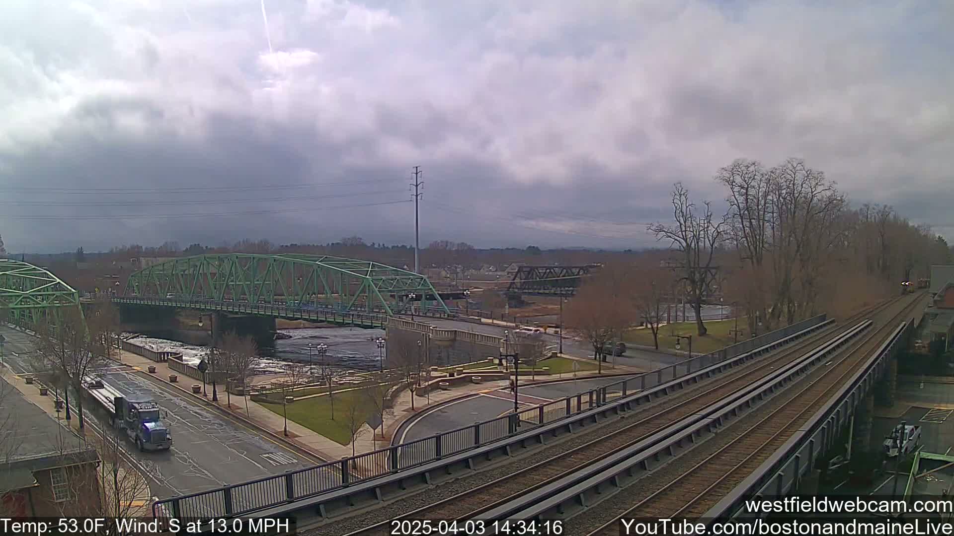 A view of two bridges over a river, a roadway, and train tracks under an overcast sky with a south wind at 13 mph.