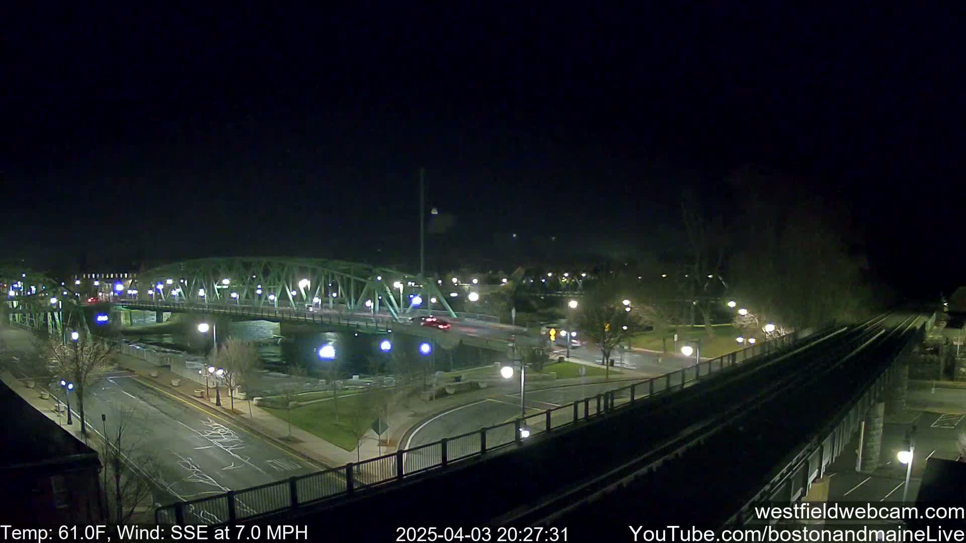 A nighttime view of a bridge over a river, with sparsely trafficked roads and a railway track visible, under a clear, dark sky.