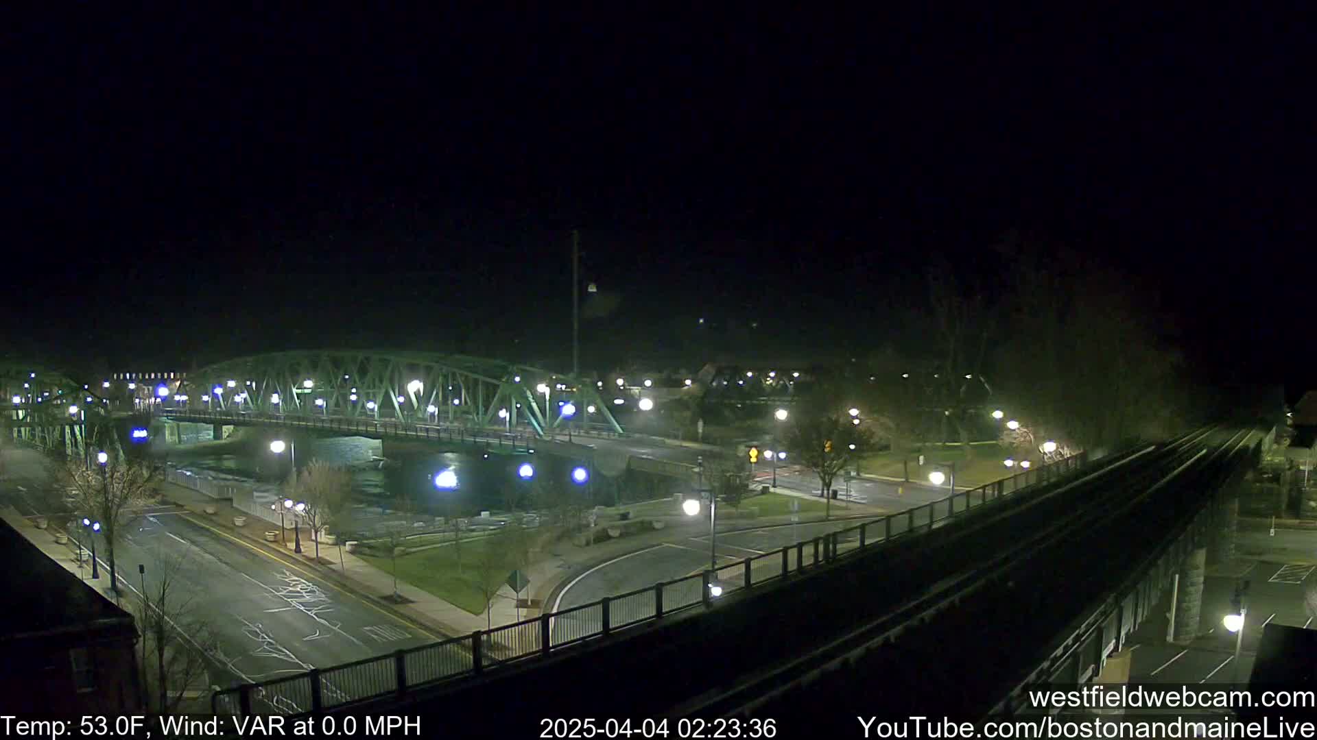 A nighttime view of a green metal bridge spanning a river, with roads and a train track visible below under a clear, dark sky.