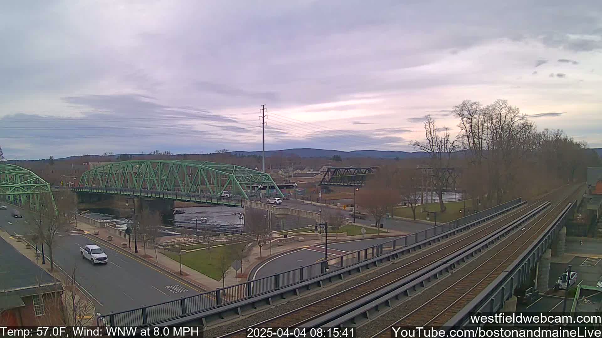 A green bridge spans a river, with another bridge and train tracks visible, under a partly cloudy sky.