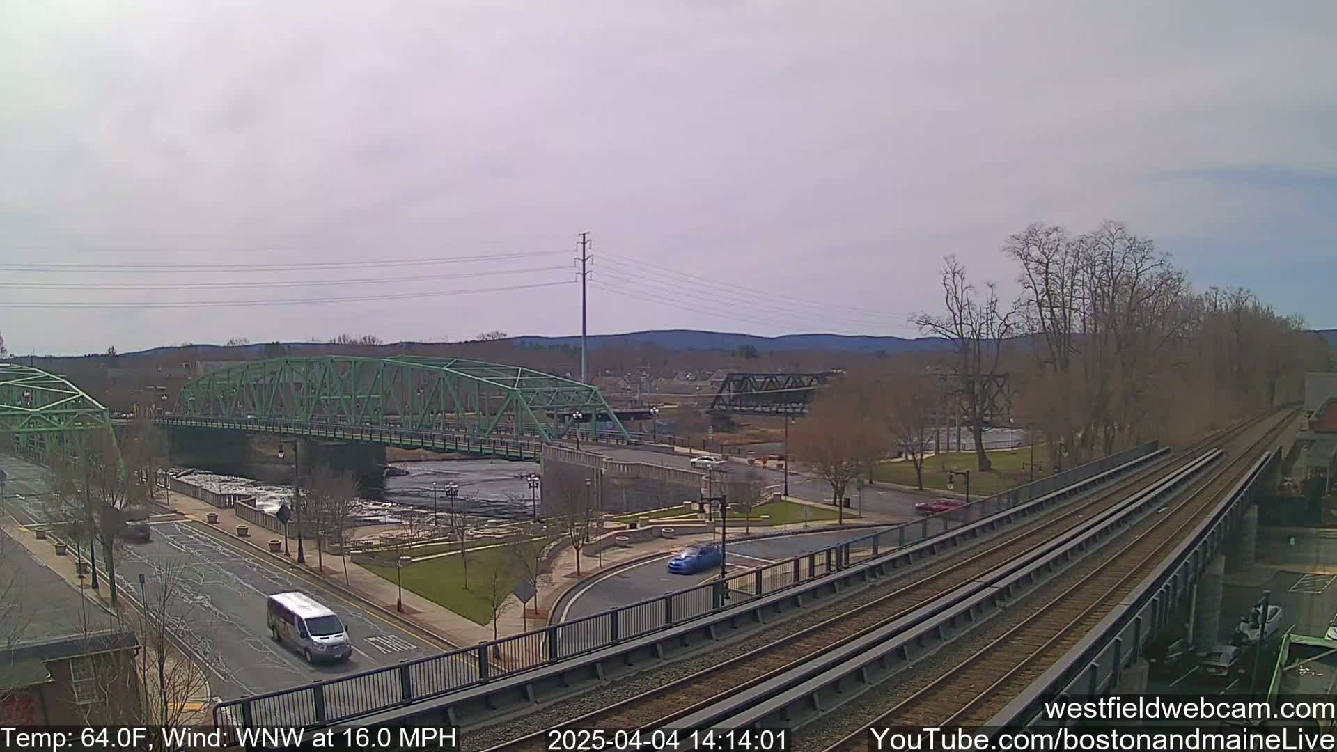 A view of a river with two bridges, one green and one dark gray, under a cloudy sky with a van and some cars visible on the roads near a train track.